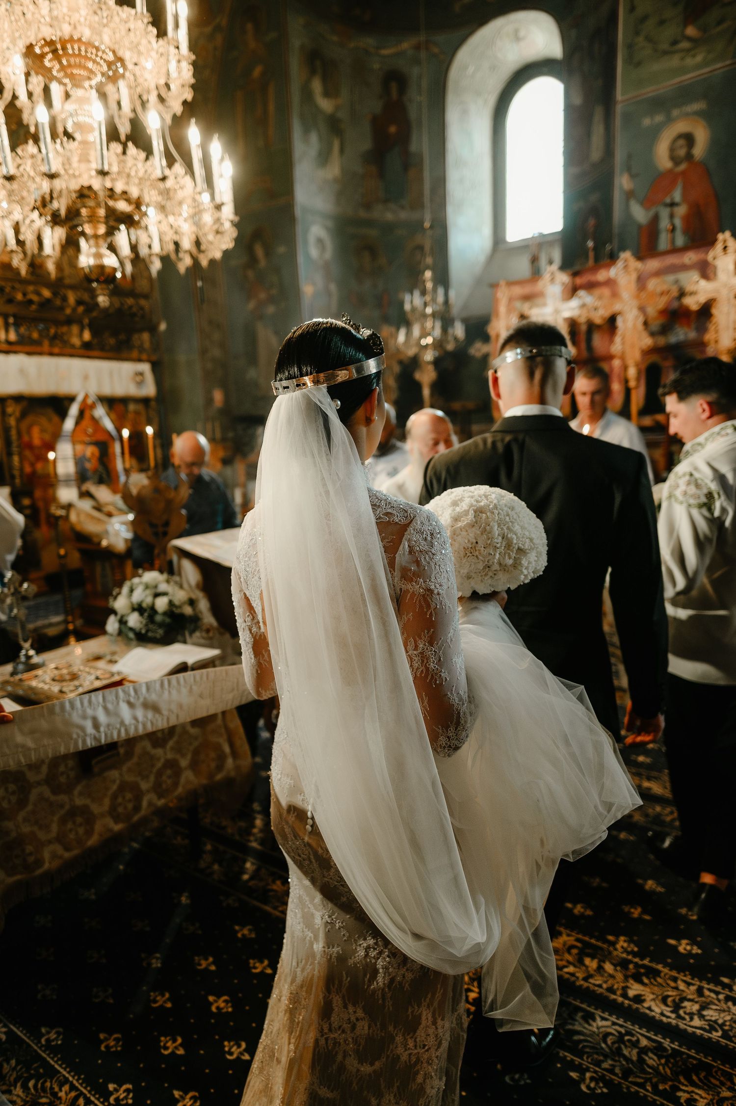 Bride in lace gown with crown and veil stands at altar in decorated Orthodox church.