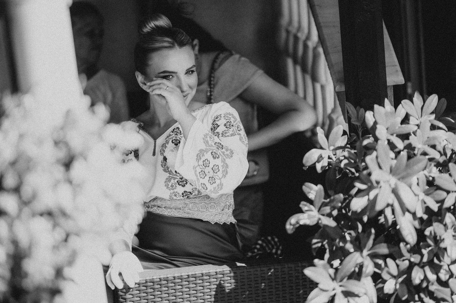 Black and white photo of woman in embroidered blouse seated near blooming flowers.