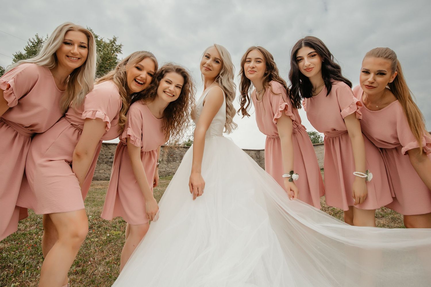Bride in white gown poses with six bridesmaids in matching dusty rose dresses outdoors on wedding day.