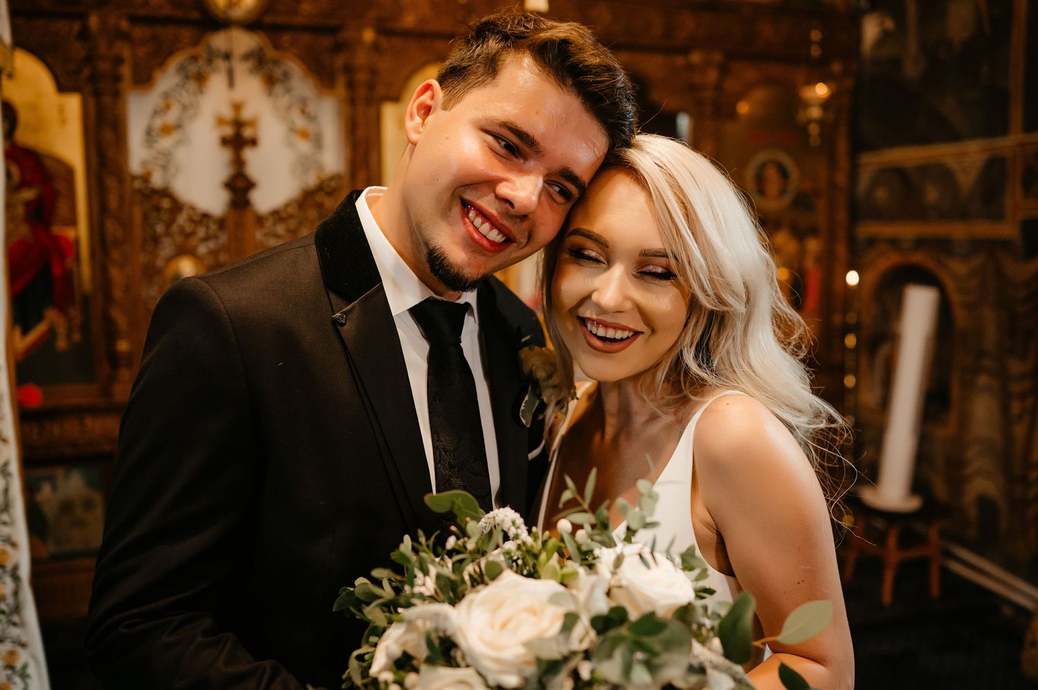 Smiling bride and groom embracing with white floral bouquet after wedding ceremony.