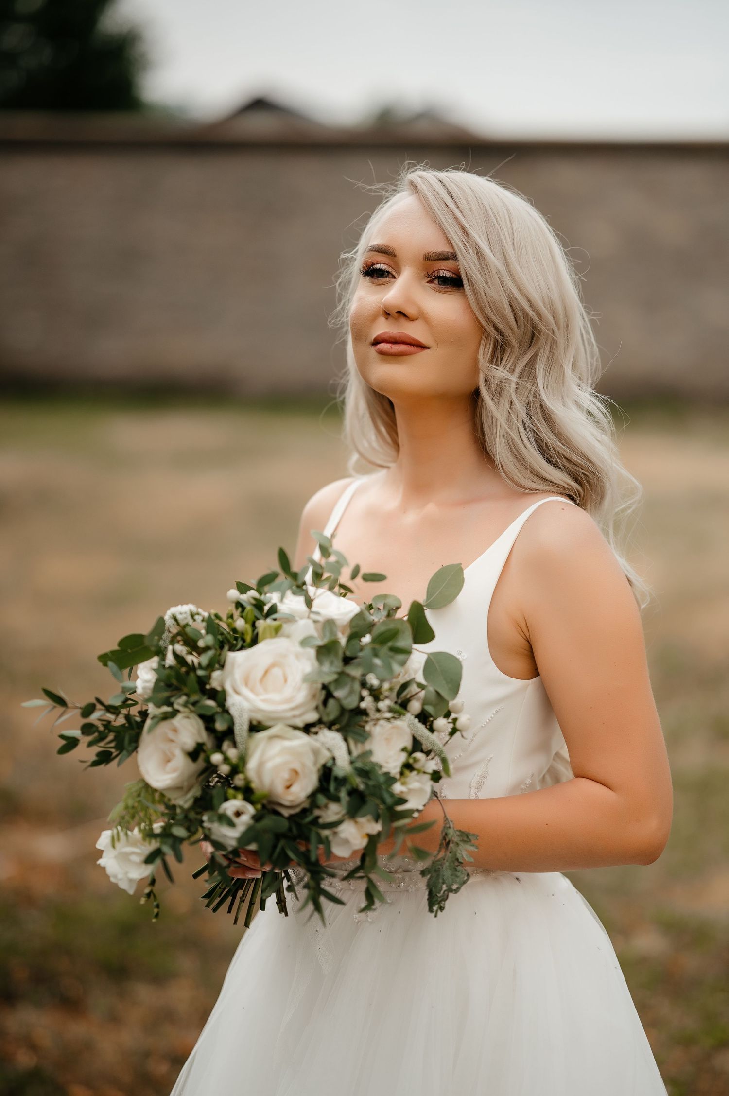Elegant blonde bride holding a white rose and eucalyptus bouquet poses in soft outdoor light.