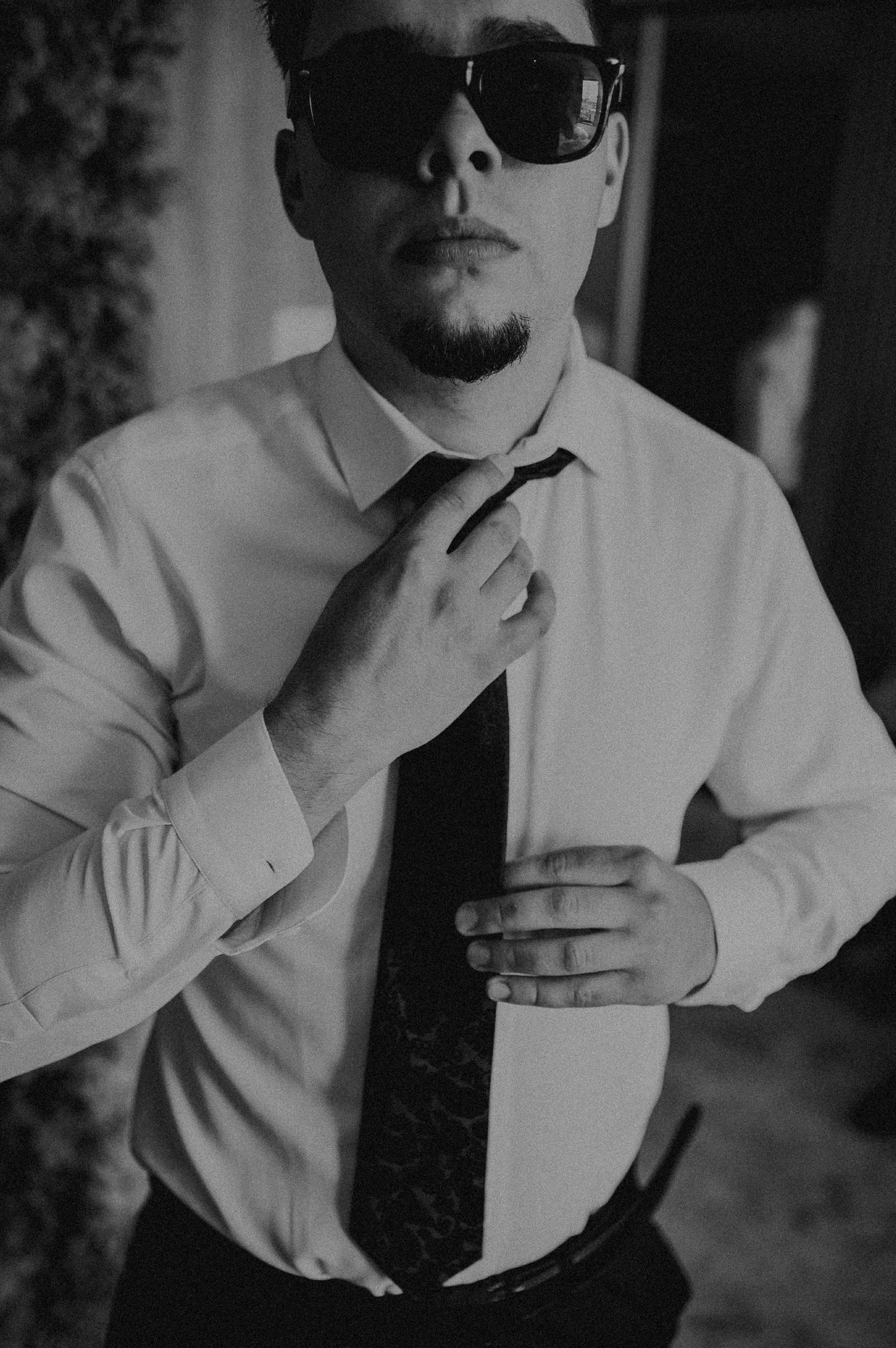 Black and white portrait of groom in white shirt adjusting tie while wearing stylish sunglasses indoors.