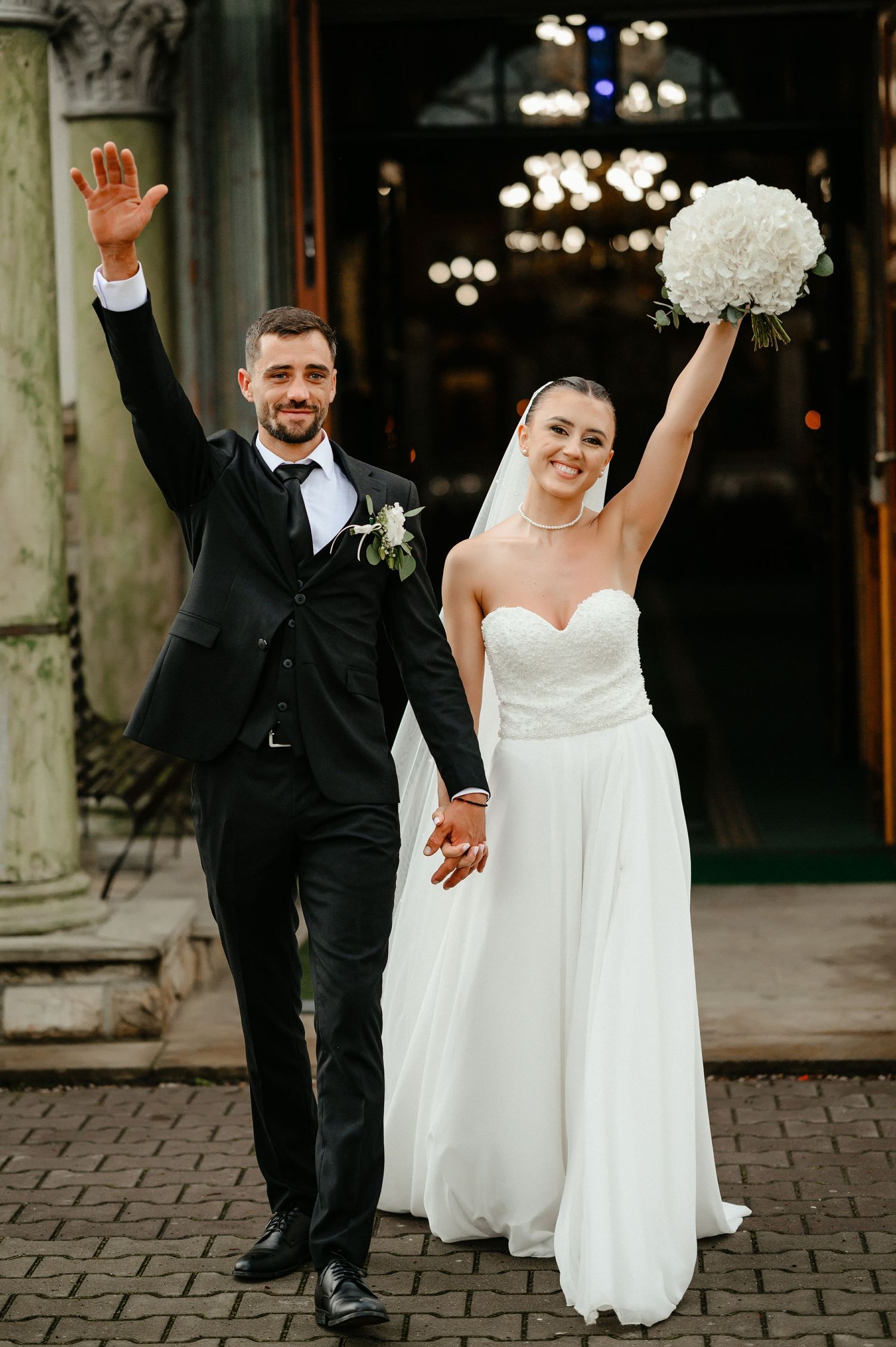 Joyful newlyweds celebrating outside church, bride in white gown raising bouquet, groom in black suit waving.