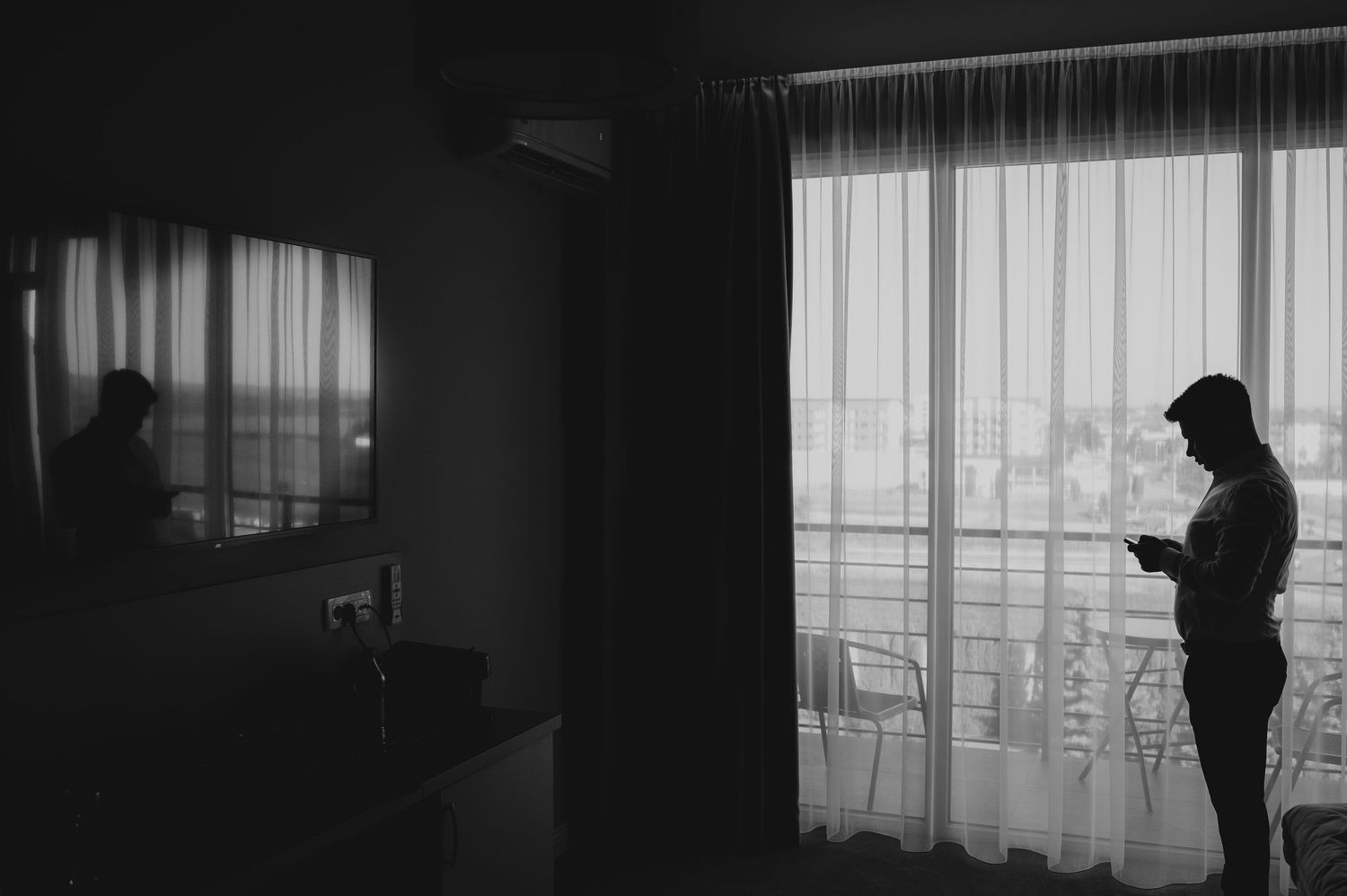Black and white silhouette of groom standing by sheer curtained window in dark hotel room.