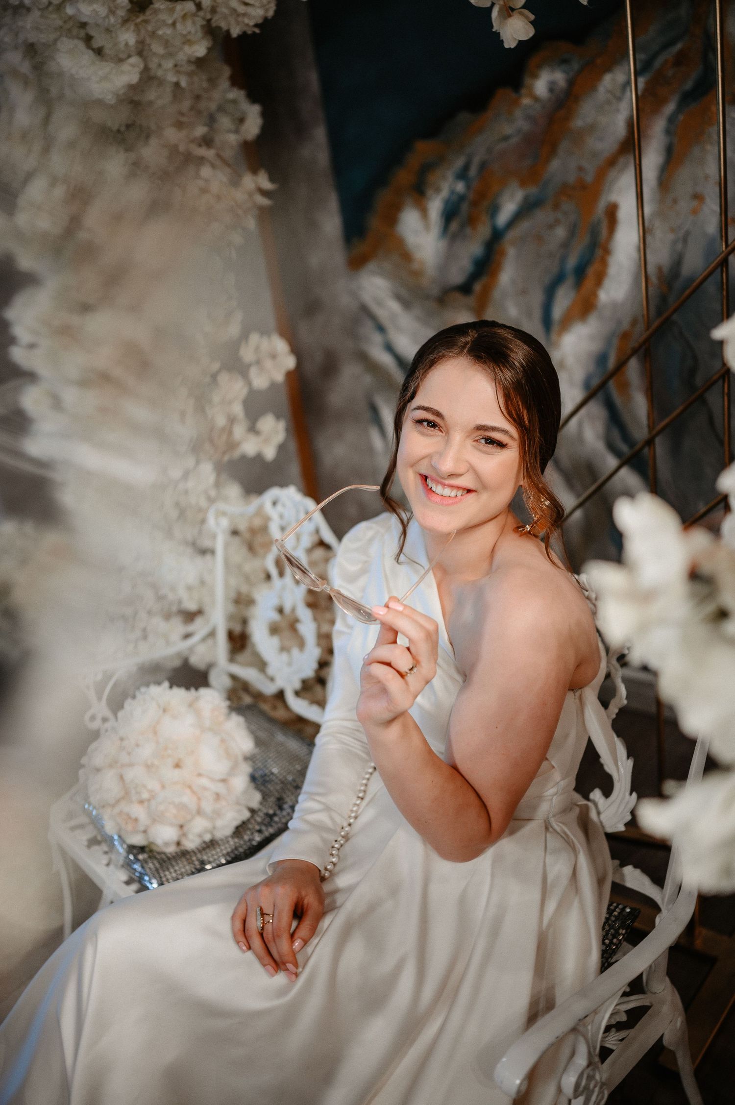 Smiling bride in white strapless gown sits beside white rose bouquet among elegant floral decorations.
