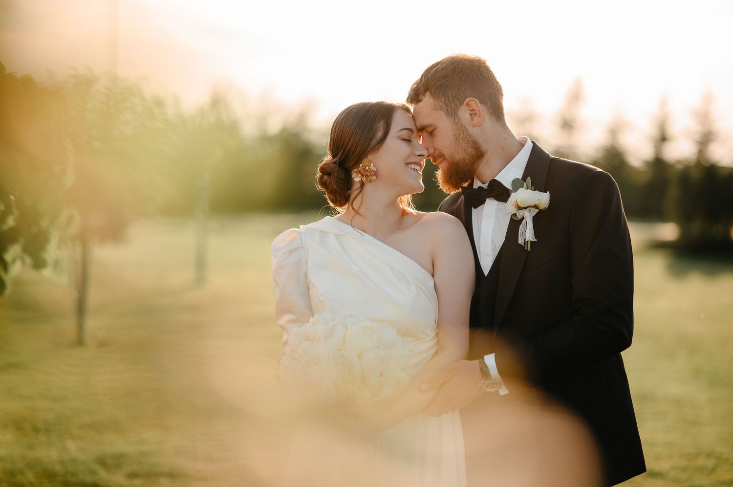 Couple shares an intimate close moment during golden hour sunset, groom in tuxedo and bride in white gown.