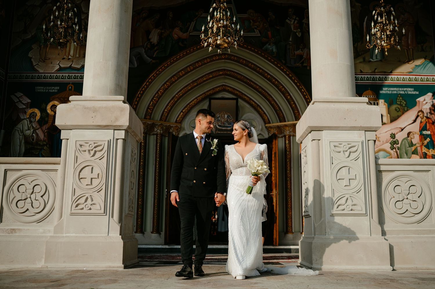 Elegant bride and groom posing at ornate Orthodox church entrance with decorative stone columns and arch.