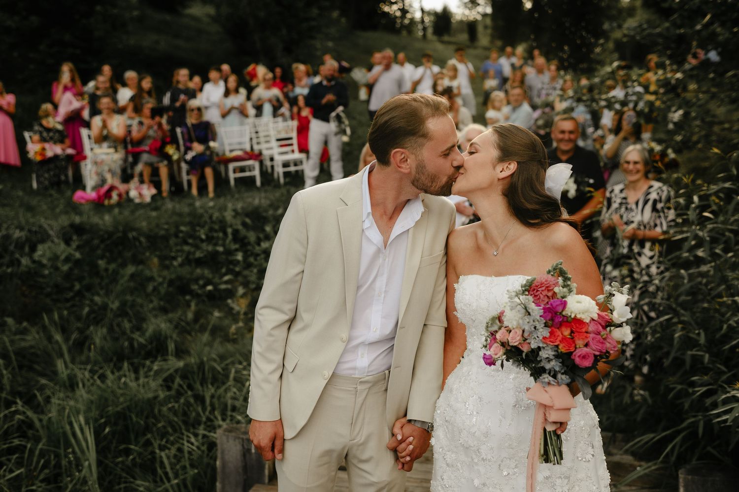 Groom kisses bride holding vibrant bouquet while guests celebrate in background at outdoor wedding.