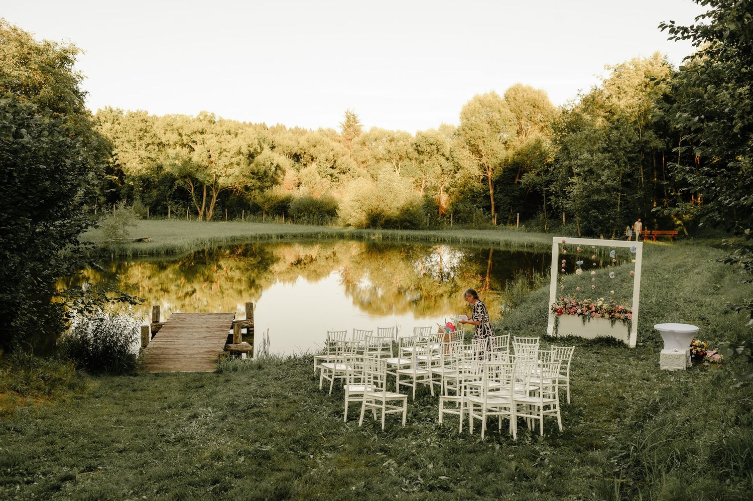 Outdoor wedding ceremony setup with white chairs beside a serene pond surrounded by lush greenery.