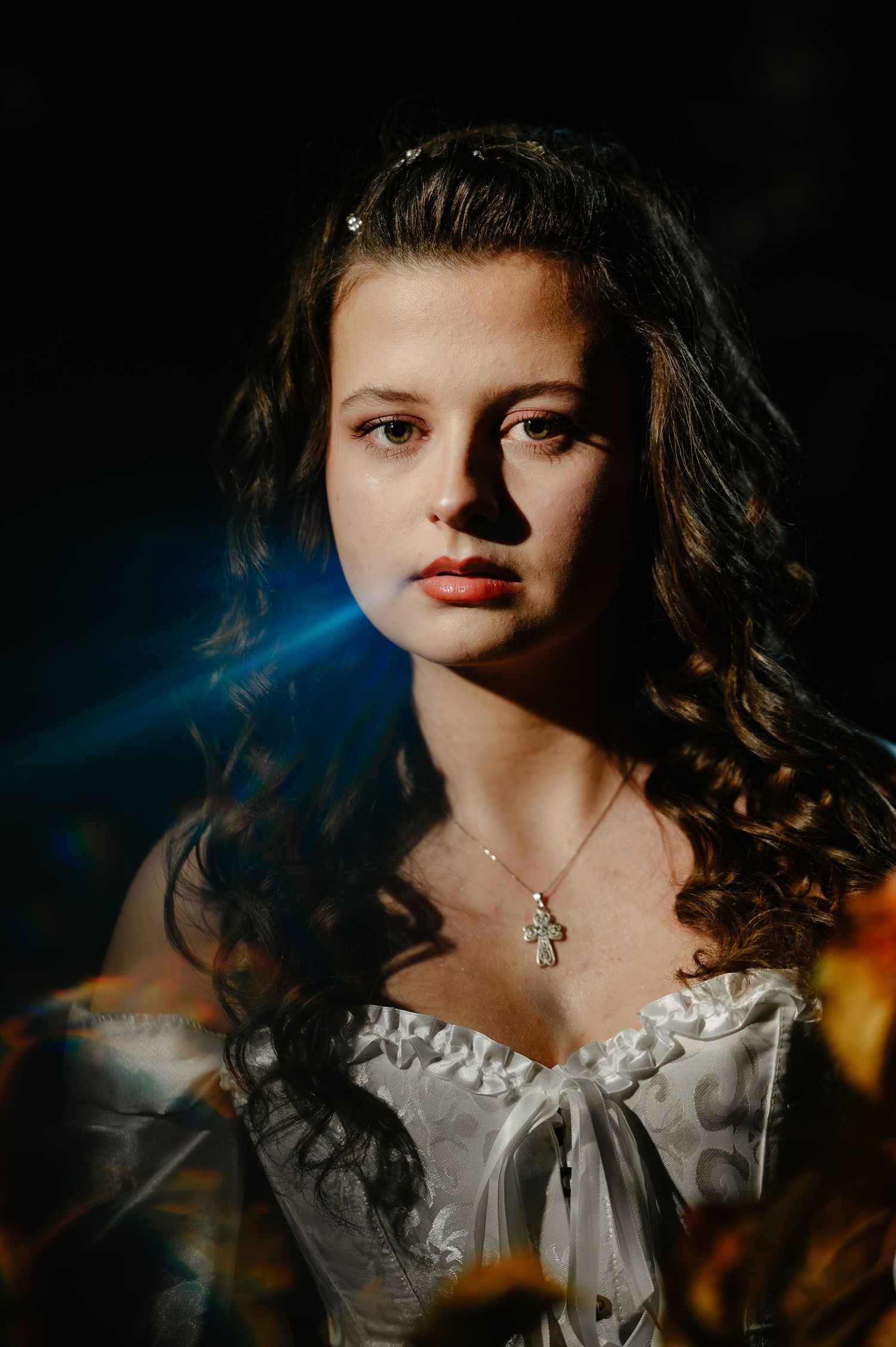 Portrait of young woman in white corset dress with cross necklace illuminated by dramatic blue light at night.