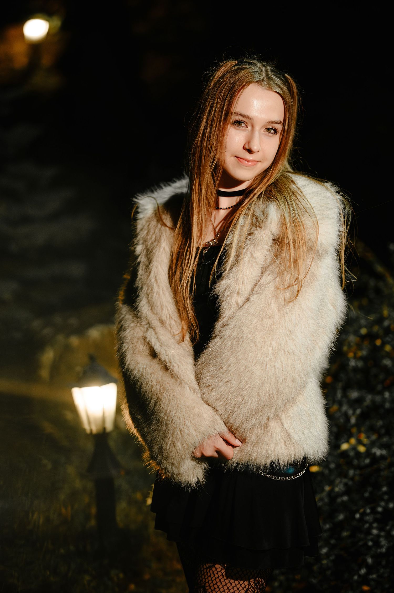 Young woman in fluffy white fur coat poses outdoors at night near a glowing street lamp.