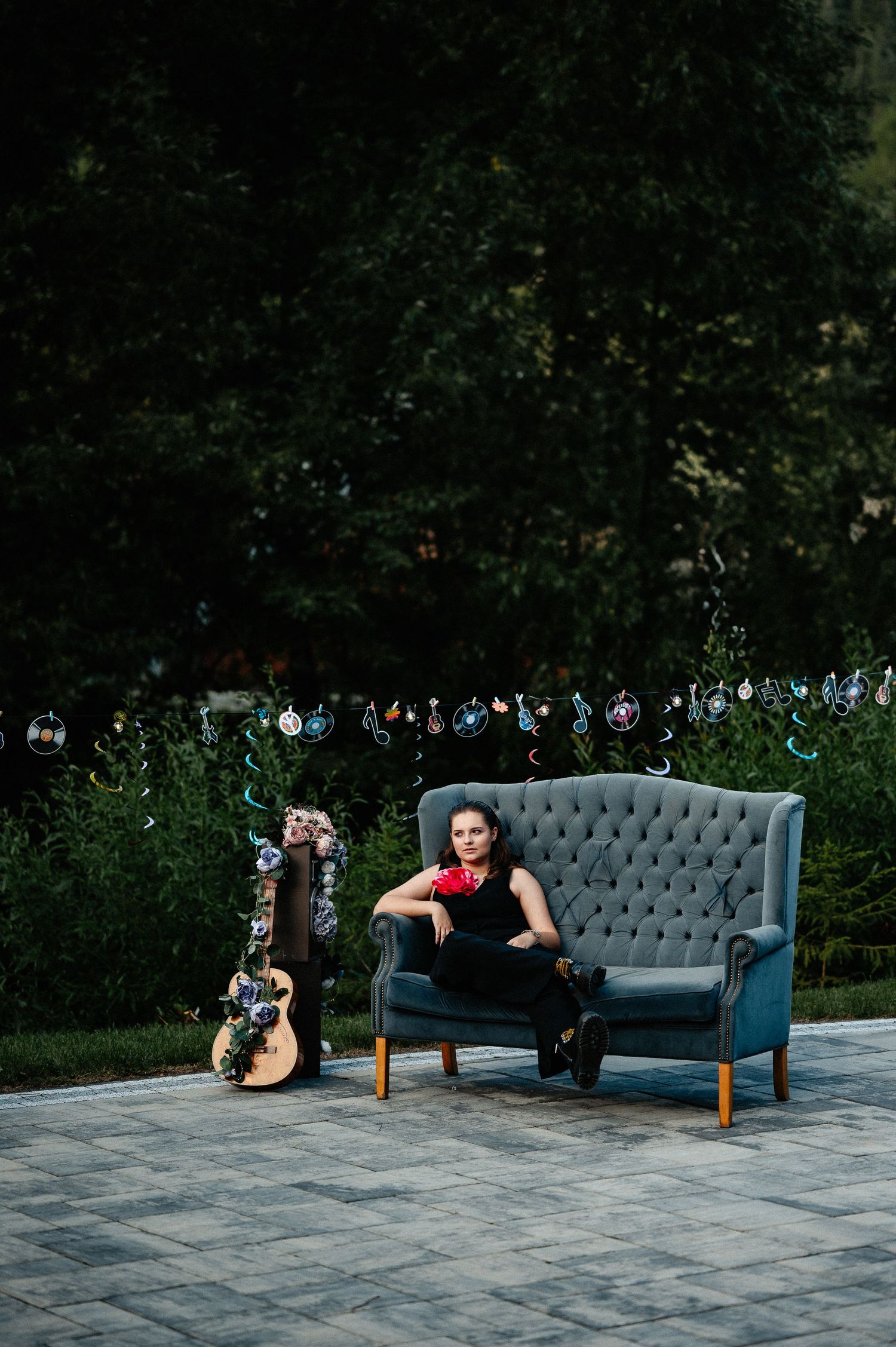 Young woman in black outfit with red corsage sitting on gray velvet sofa beside a decorated guitar.
