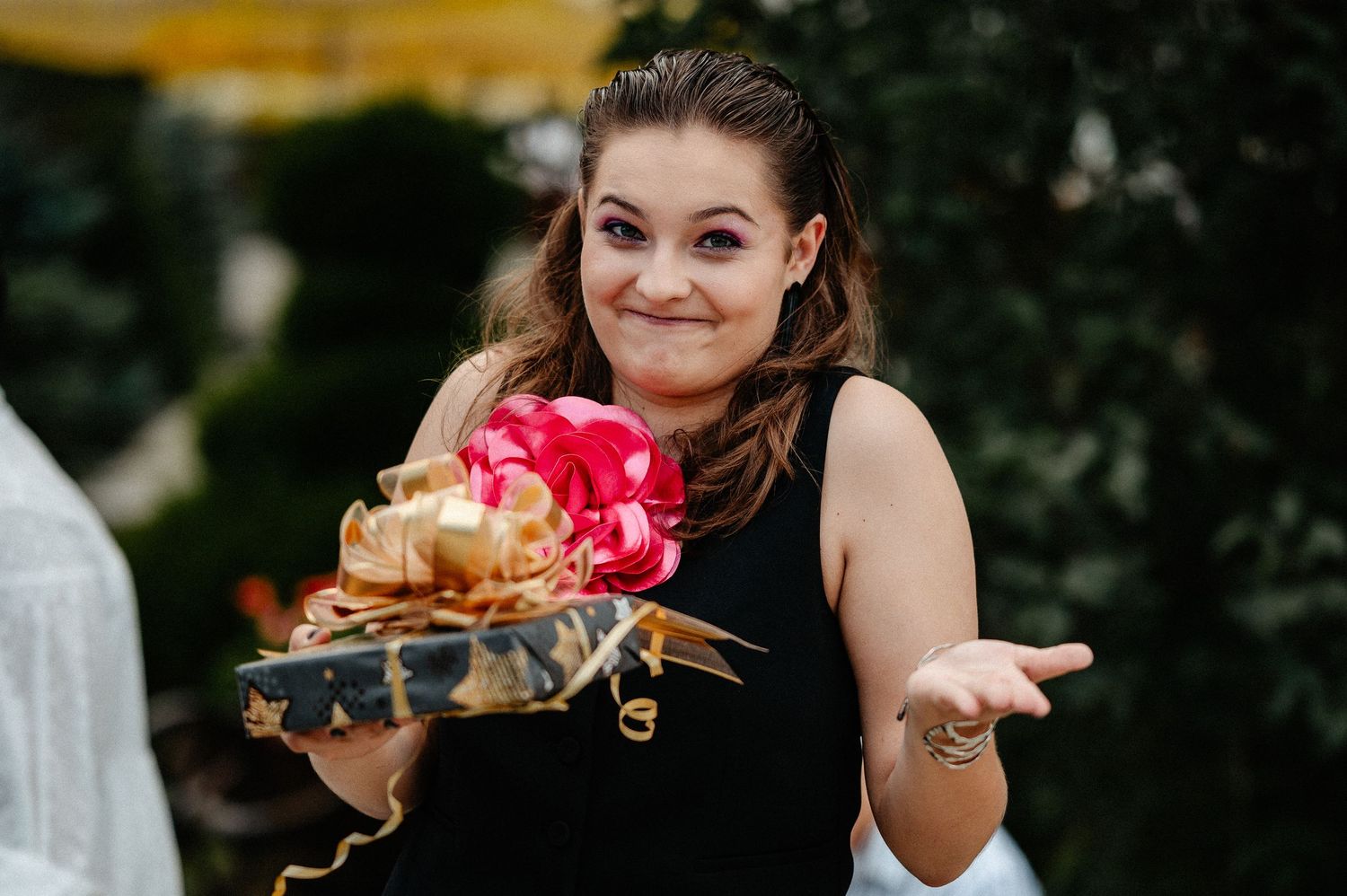 Young woman in black dress smiling while holding a gift with gold bow and red flower decoration.