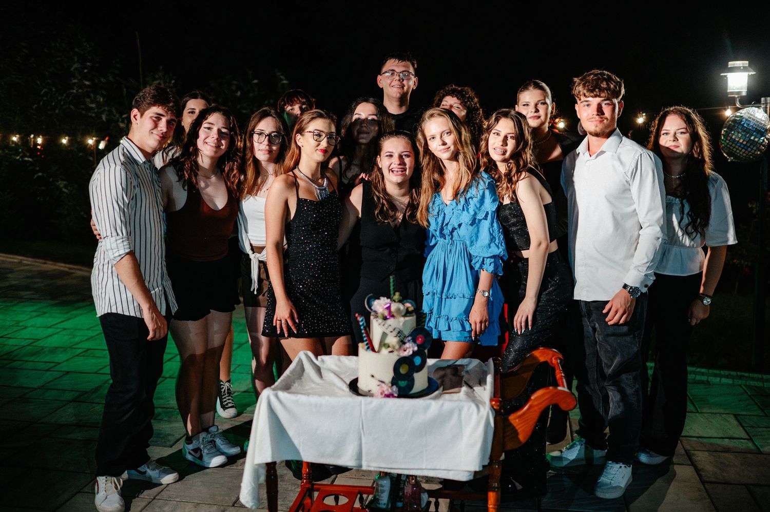Group of teenagers posing together at an outdoor night birthday party with a decorated cake on a table.