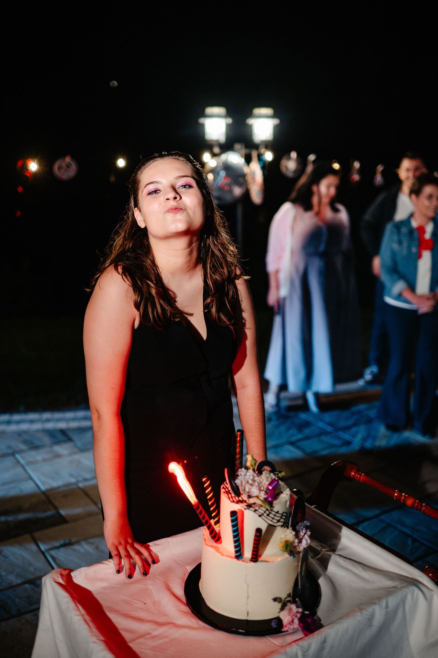 Young woman in black dress blowing out candles on white birthday cake at outdoor night celebration.