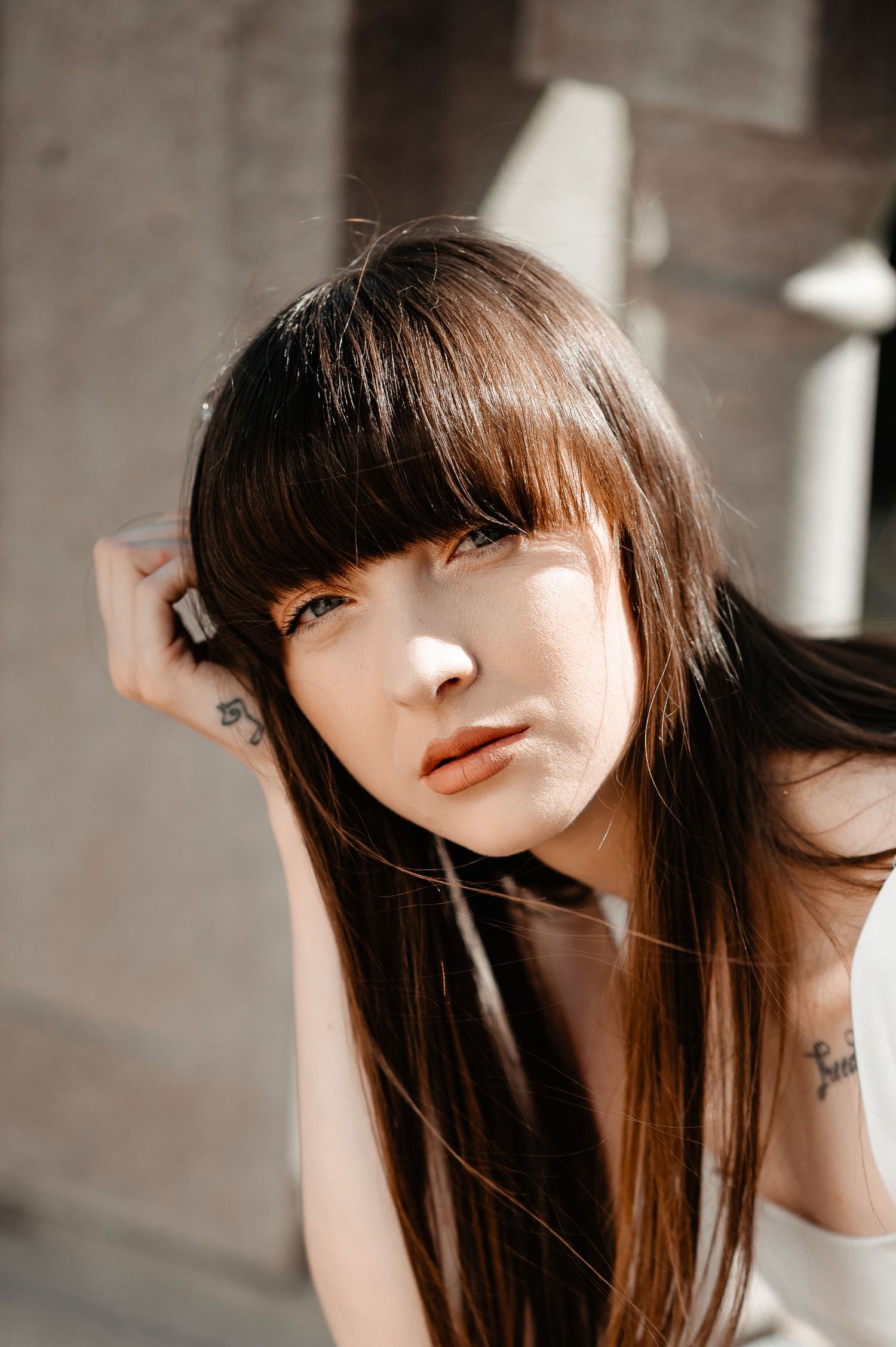 Close-up portrait of a young brunette woman with bangs and tattoos posing in soft natural light.