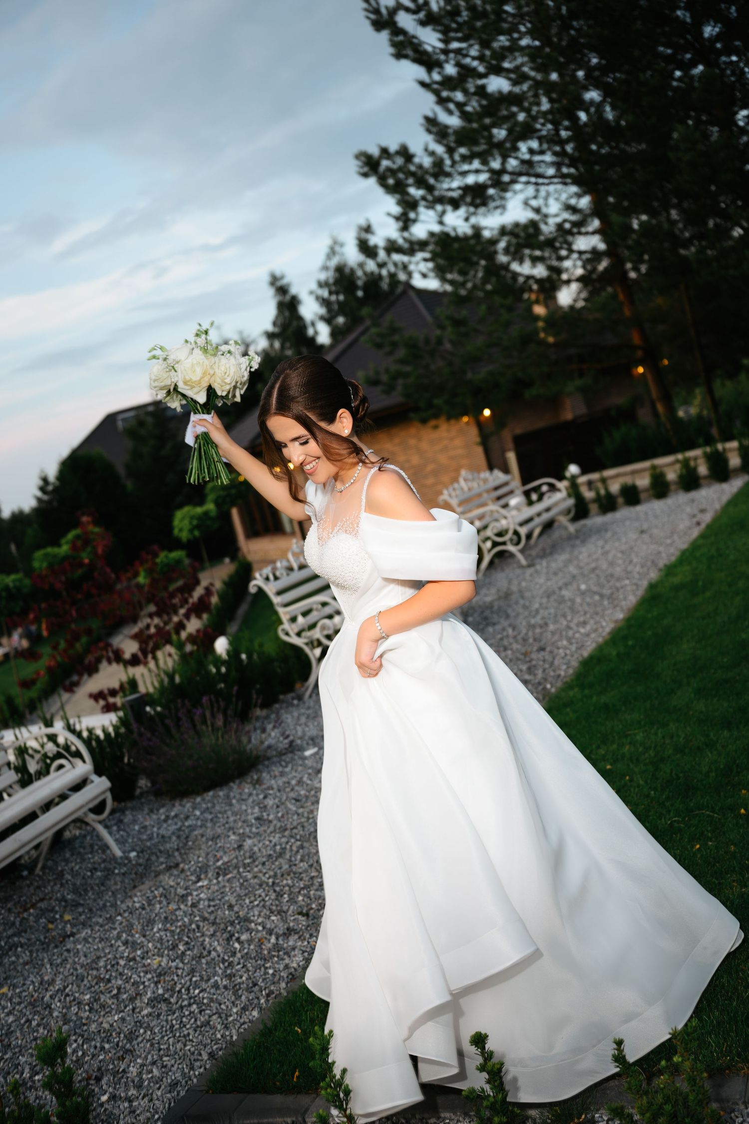 Bride in white off-shoulder gown twirling with bouquet in garden at dusk.