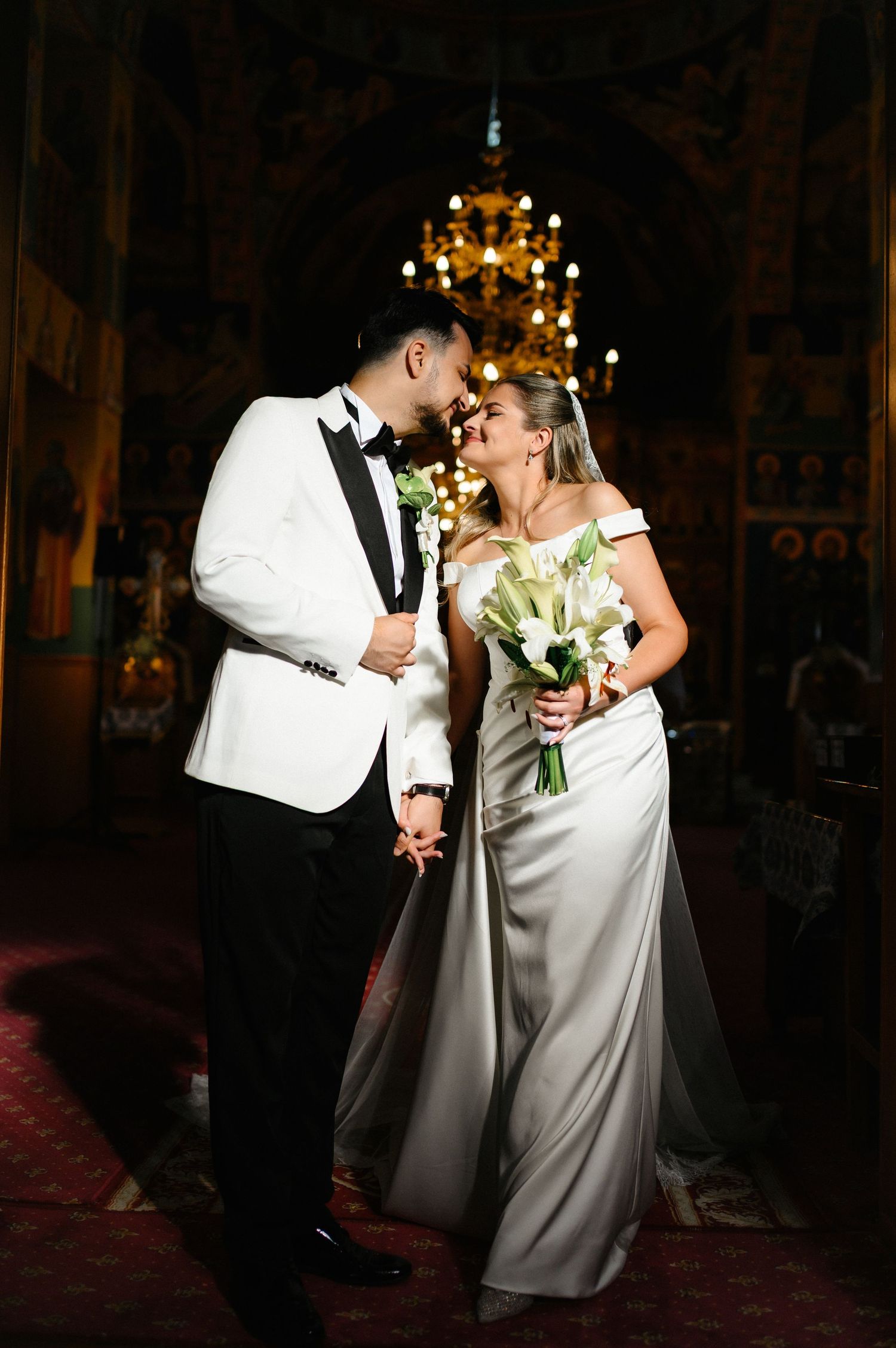Bride and groom share a romantic moment in an ornate church, bride holding white calla lily bouquet in elegant satin gown.