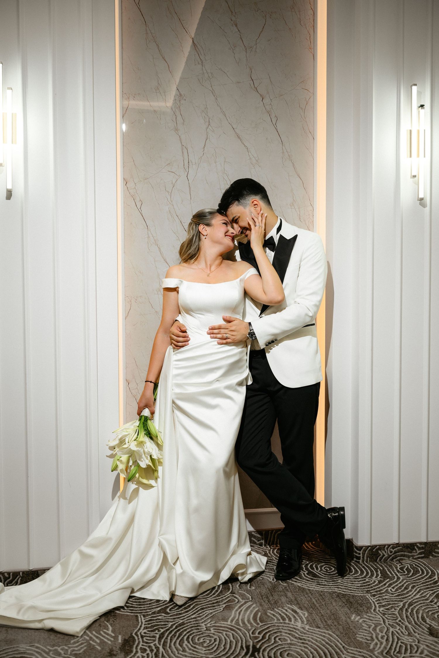 A bride in a white satin gown and groom in a white tuxedo share a romantic moment in an elegant hotel corridor.