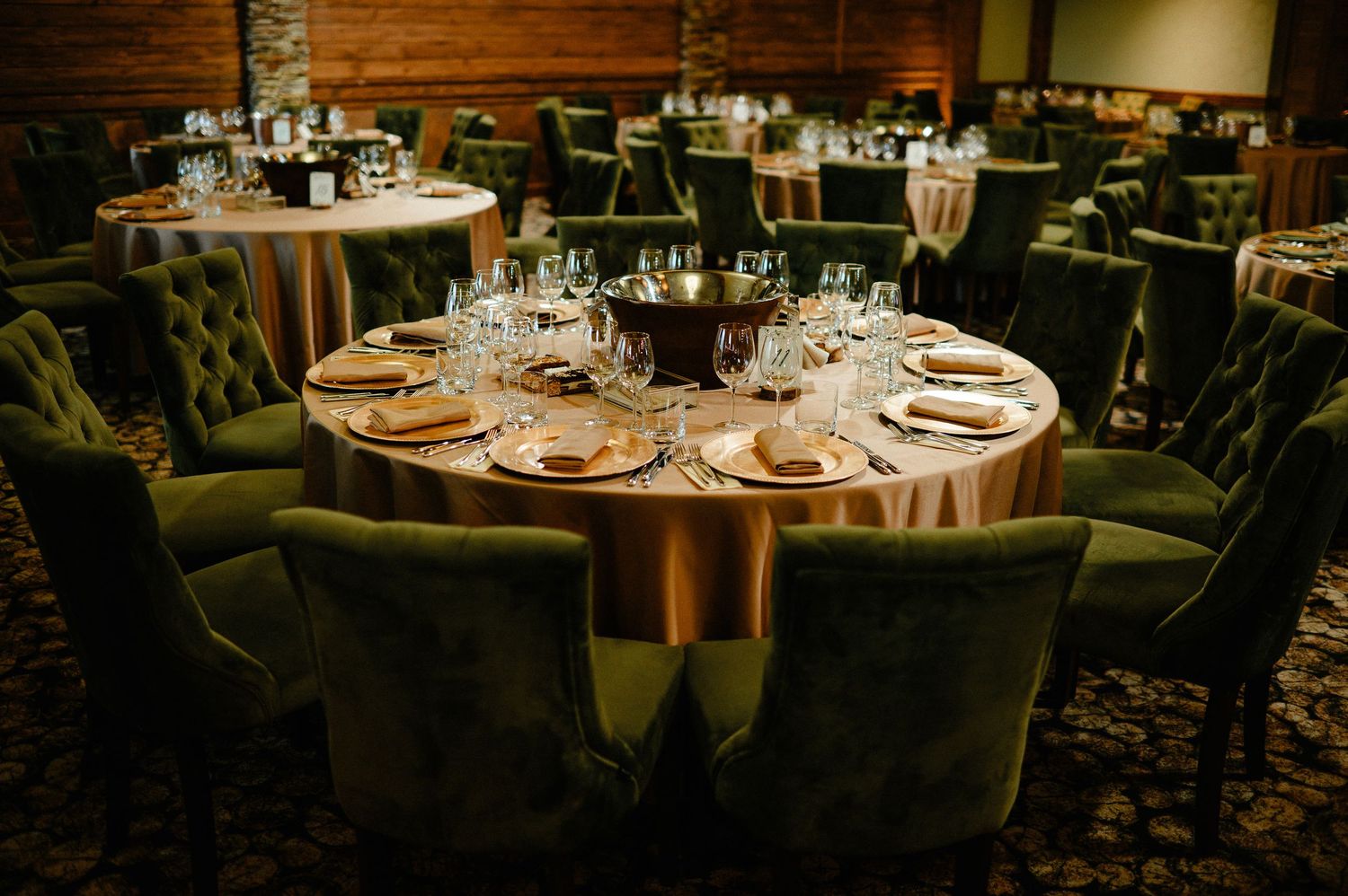 Luxurious wedding reception tables with gold linens and green tufted velvet chairs.