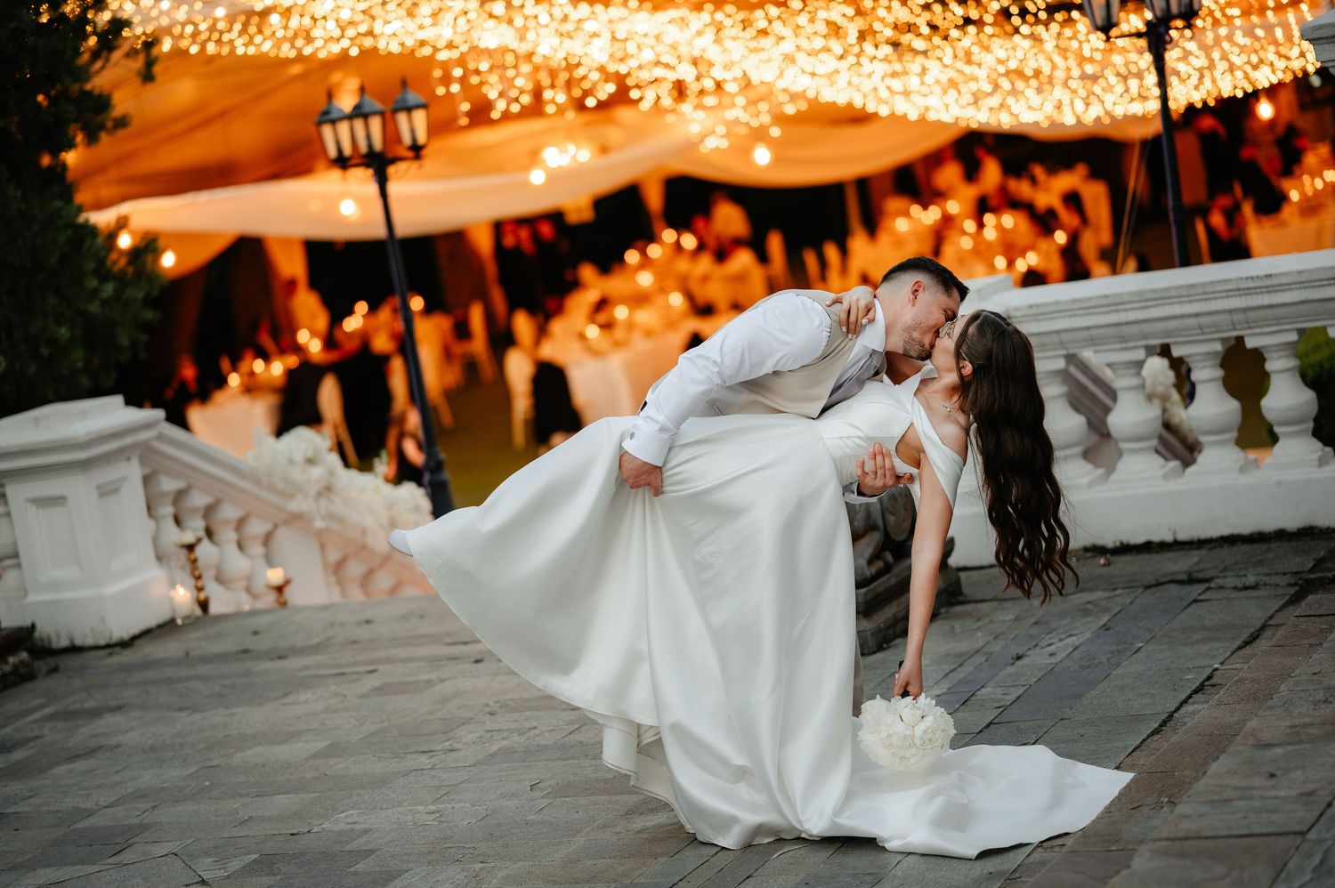 Groom dipping bride in a romantic kiss under glowing string lights at their outdoor wedding venue.