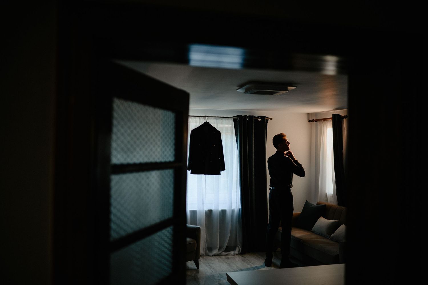 Groom standing in a dimly lit room with his suit hanging by a bright window.