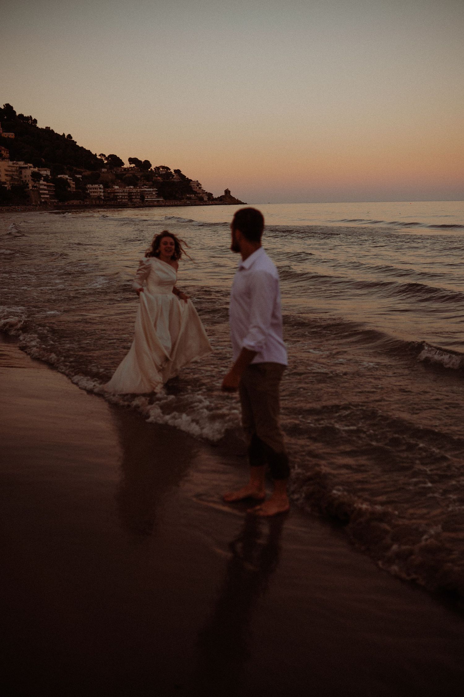 Couple in wedding attire walking along a sandy beach at sunset with ocean waves and coastal town in background.