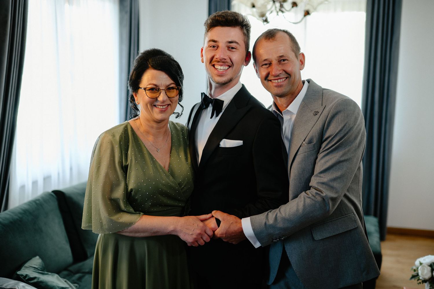 Groom in black tuxedo smiling with parents in elegant wedding attire indoors.