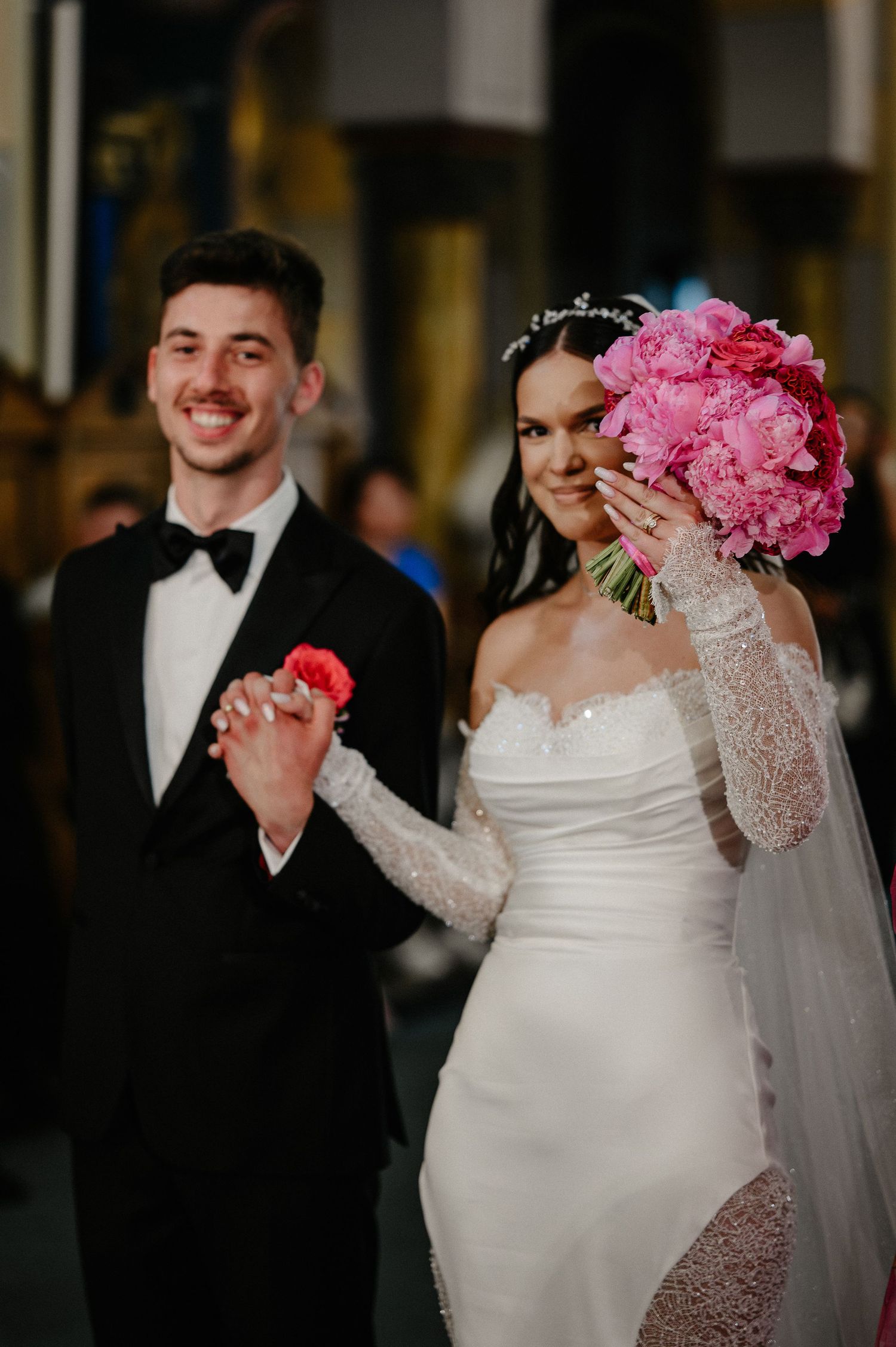 Smiling newlyweds hold hands as bride covers face with pink peony bouquet after church ceremony.