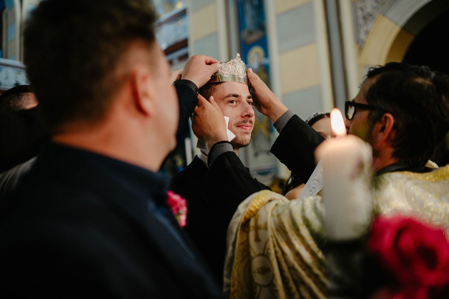 Smiling groom receives ceremonial crown placement by Orthodox priest during wedding ceremony.