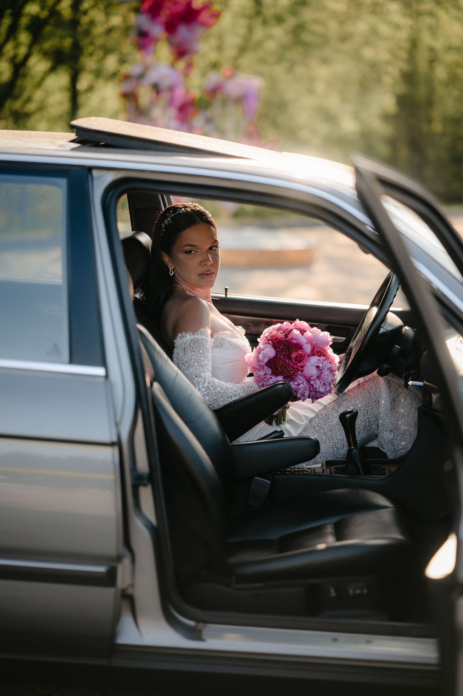 Smiling bride in lace wedding dress sits in classic car holding pink peony bouquet through open door.