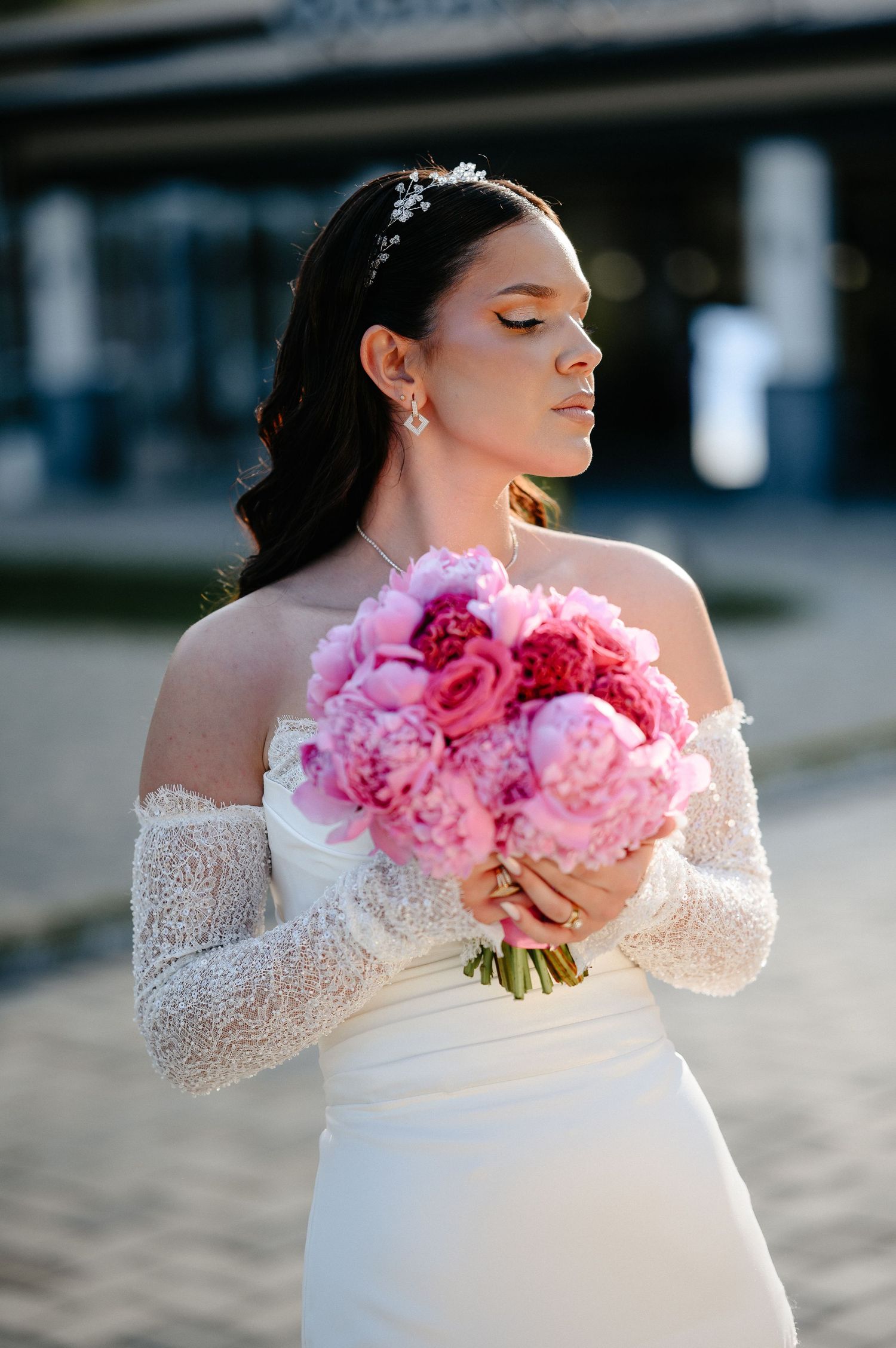 Bride with eyes closed holding vibrant pink peony bouquet, wearing white off-shoulder lace wedding gown outdoors.