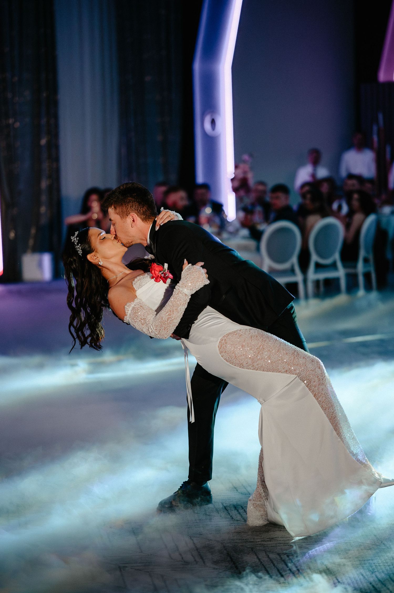 Groom dips bride in sparkling white gown during romantic first dance on fog-covered wedding dance floor.