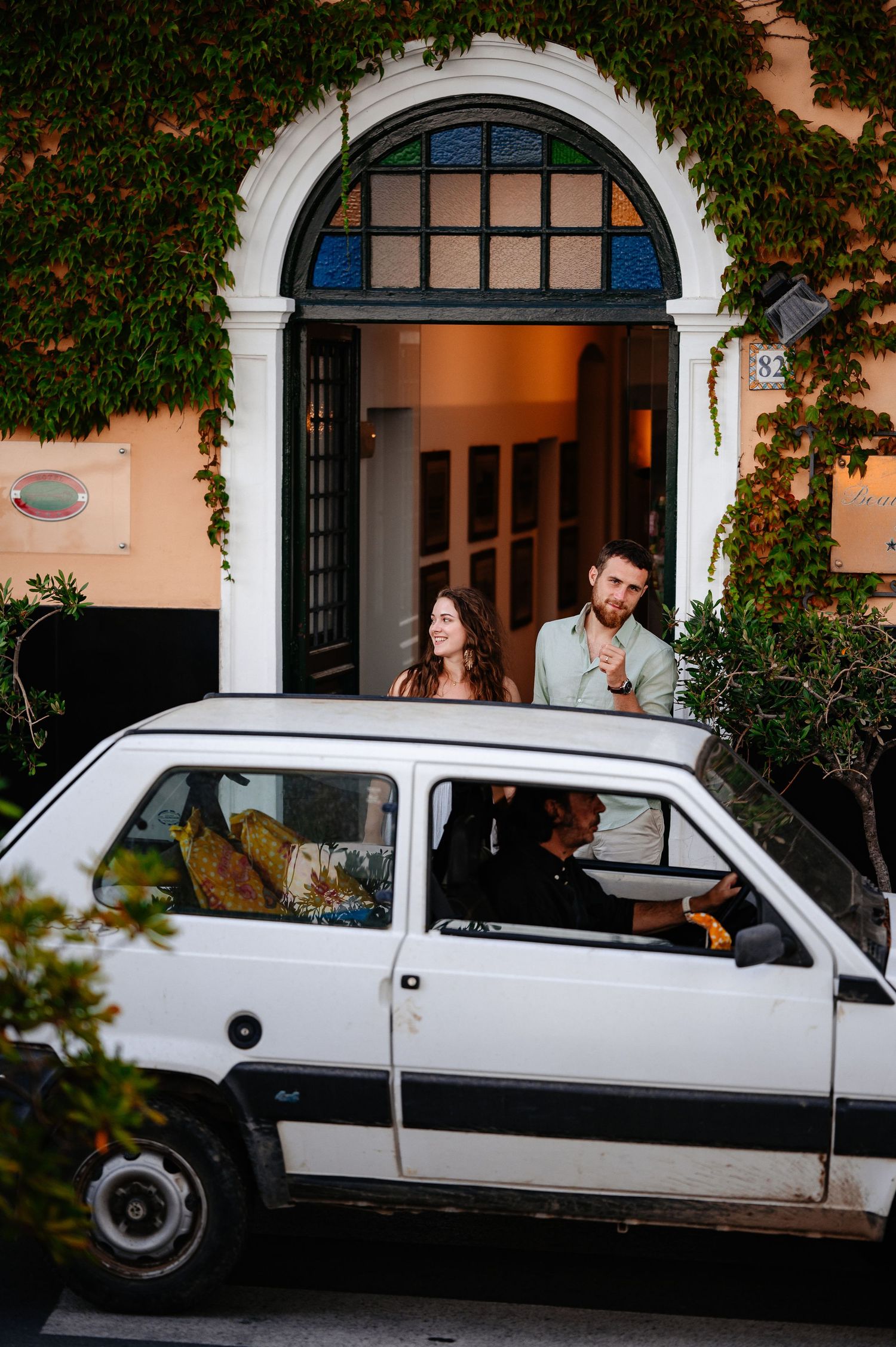 Stylish couple standing near ivy-covered building entrance beside vintage white car.