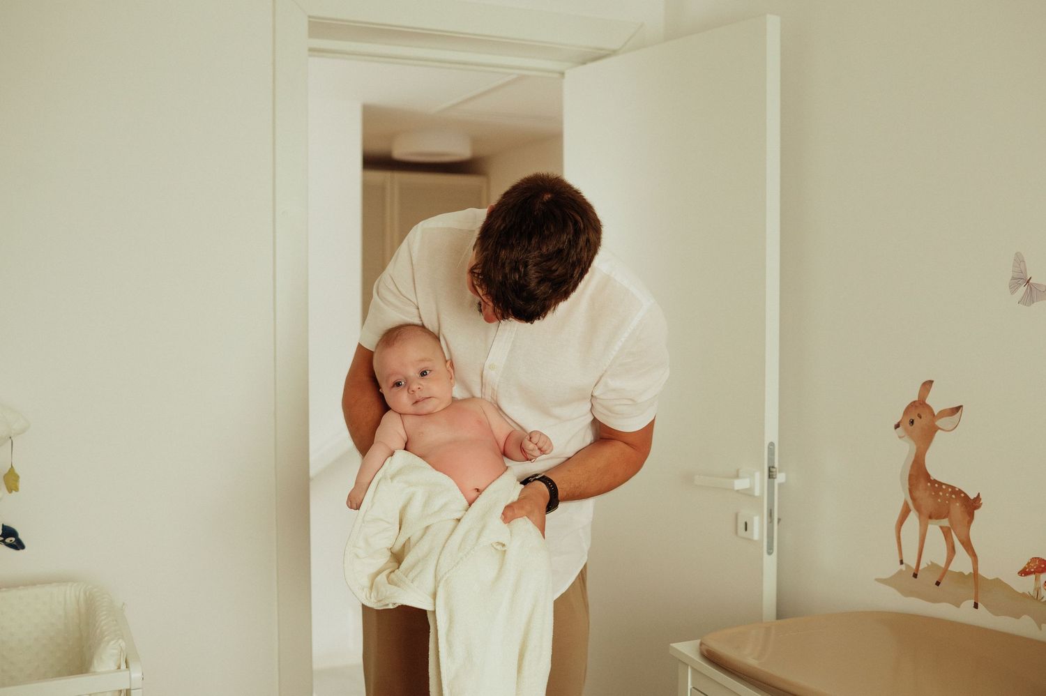 Father carefully holds newborn baby wrapped in white towel in bright nursery doorway.