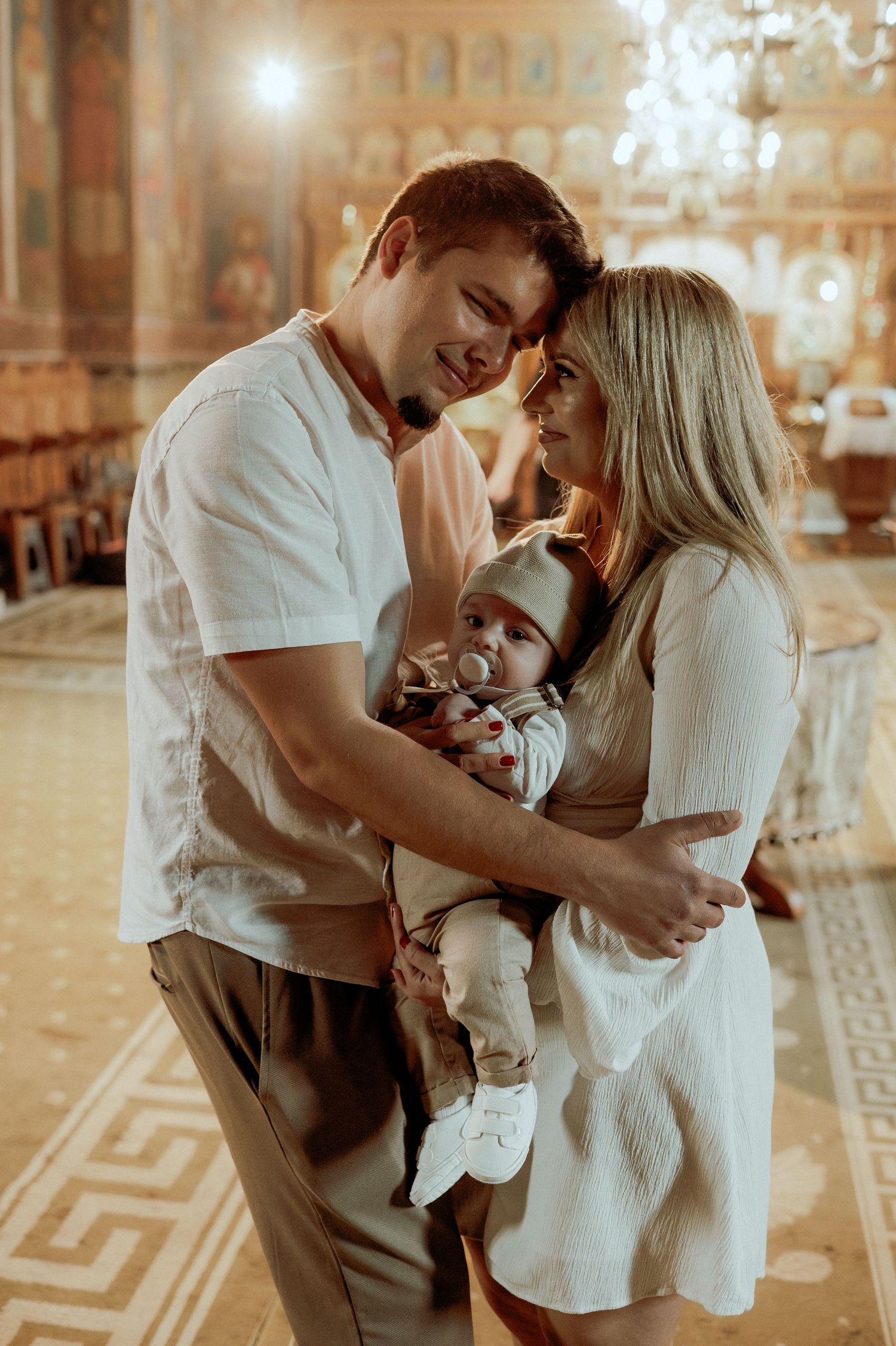 Loving couple tenderly holds newborn baby together in golden-lit Orthodox church.