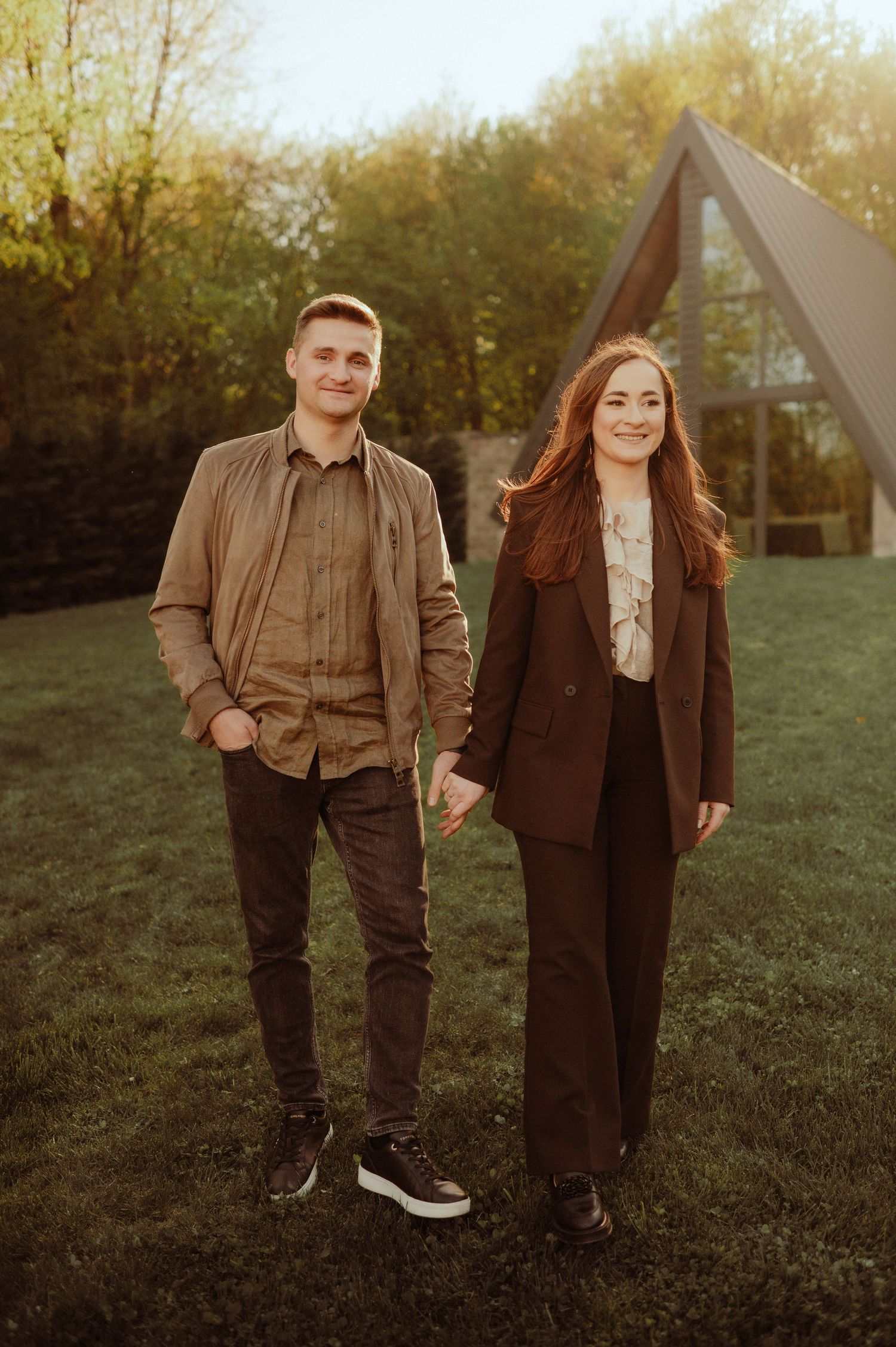 Smiling young couple holding hands outdoors in golden hour light near a modern A-frame structure.