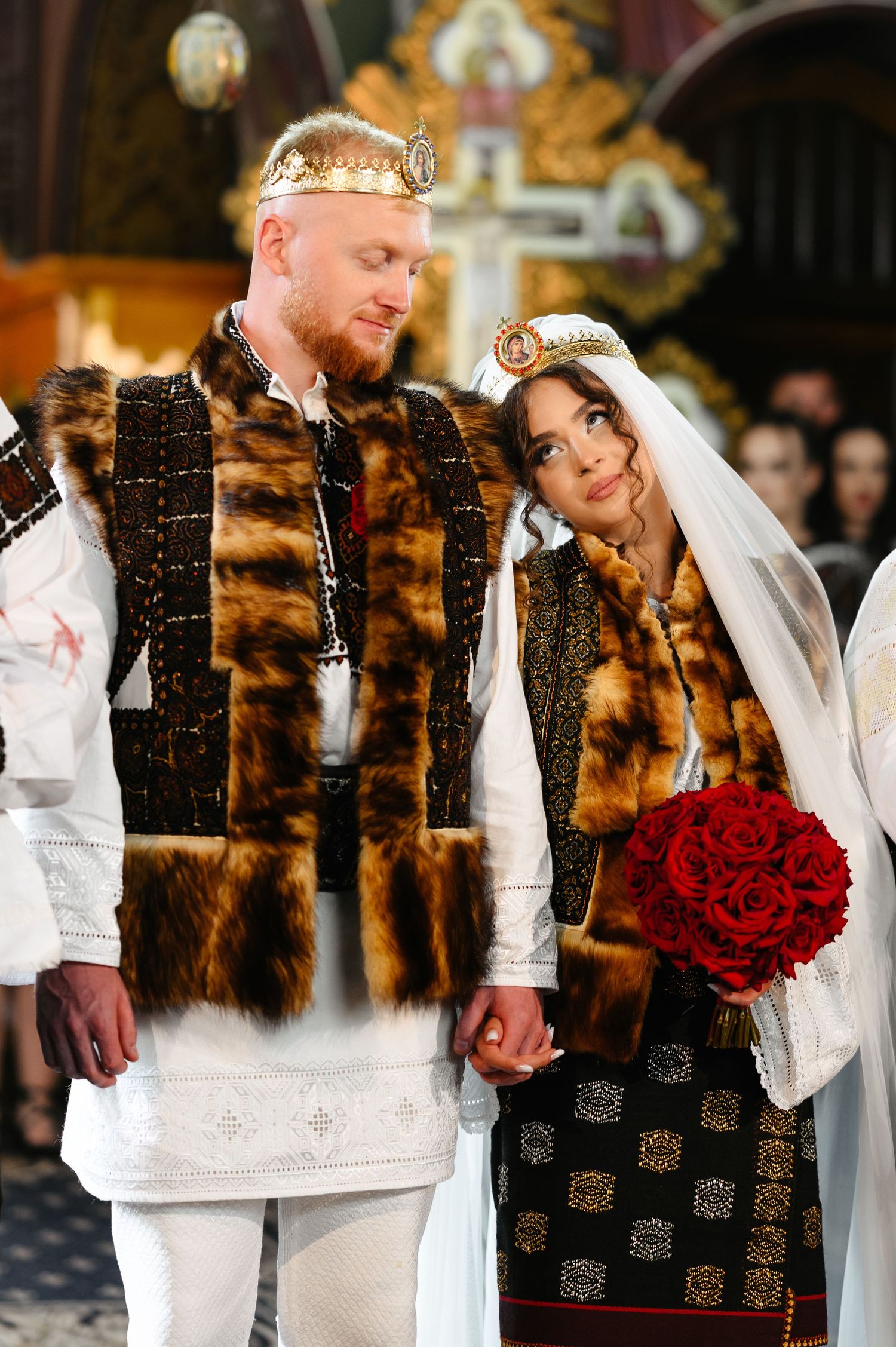 Couple in traditional Eastern European wedding attire with fur vests and crowns, bride holding red roses bouquet.