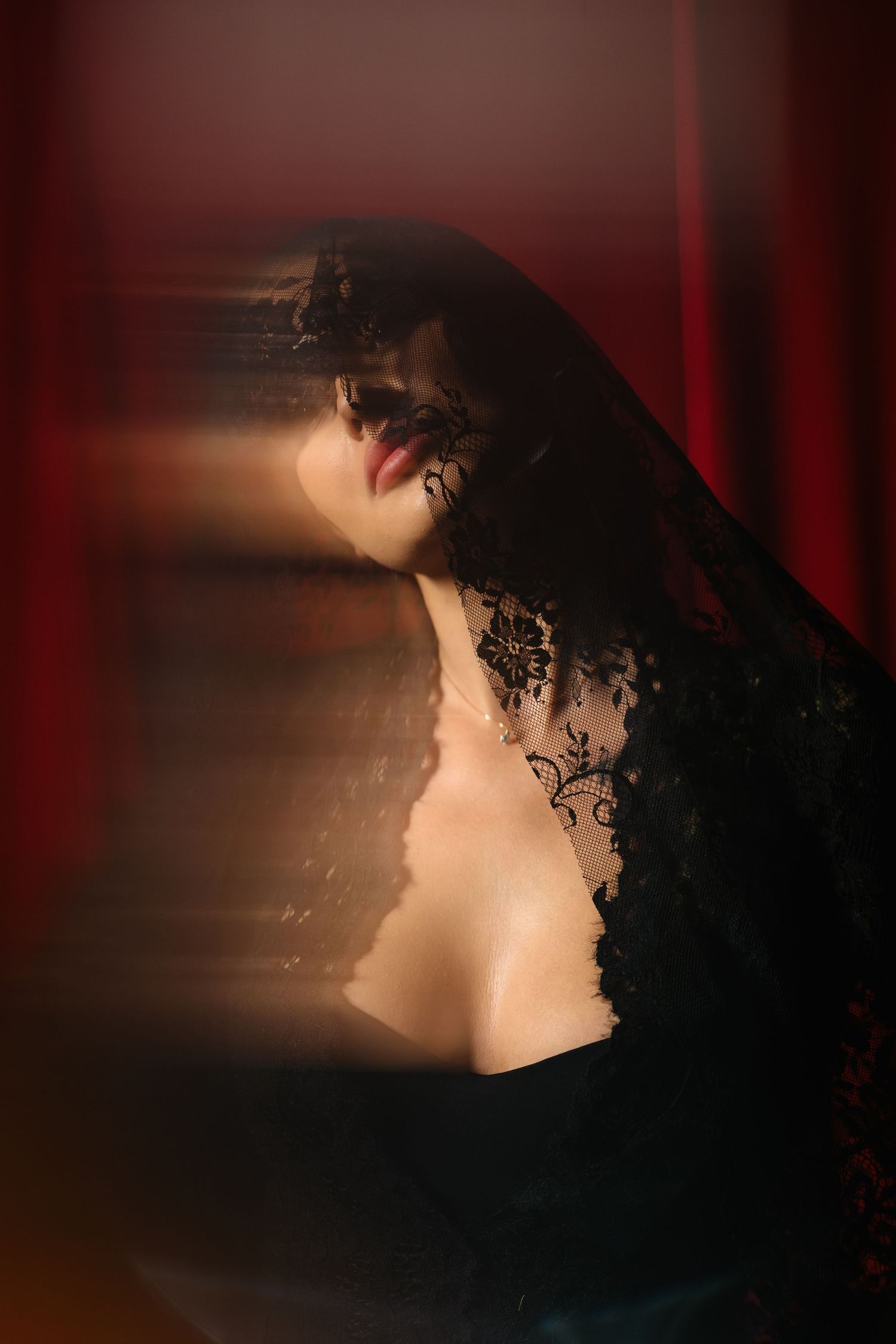A woman draped in black lace veil poses dramatically against a red background in moody, artistic portrait photography.