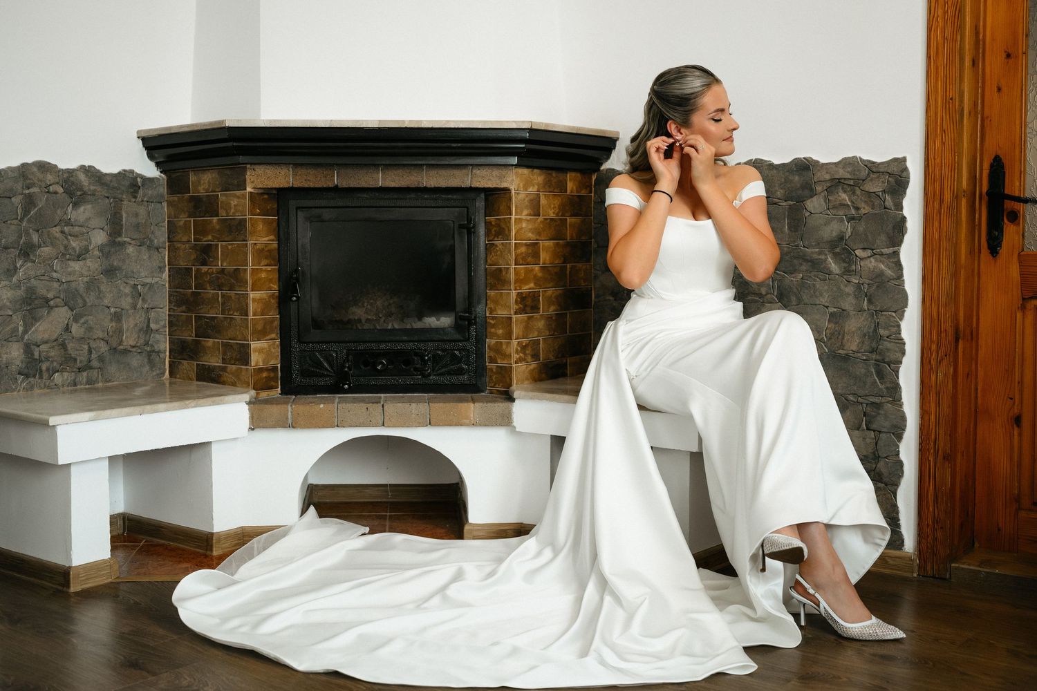 Bride in white off-shoulder gown sits by stone fireplace, adjusting earring, with elegant train and heels.