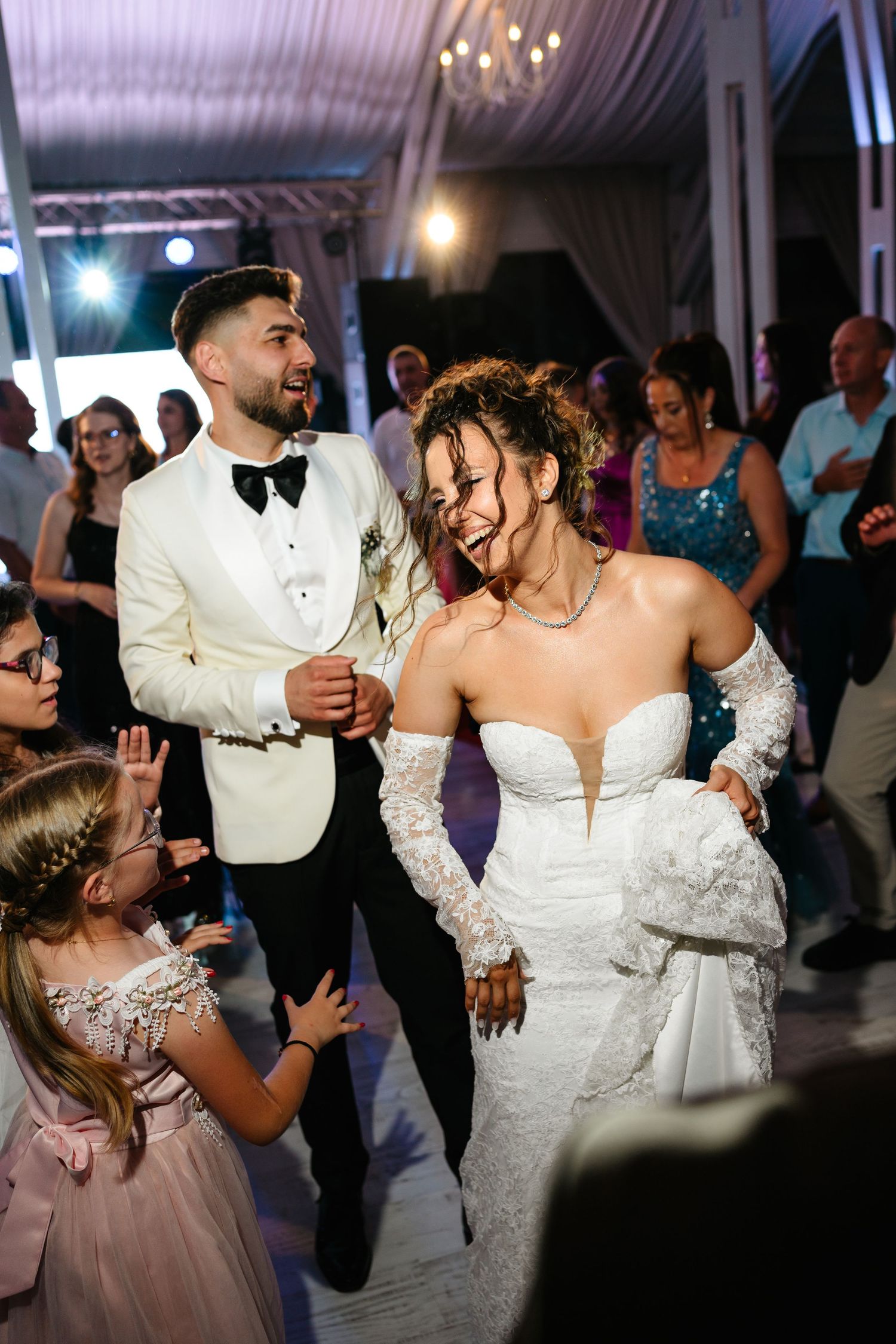 Bride and groom dancing joyfully with guests at elegant wedding reception, bride in strapless lace gown, groom in white tuxedo.