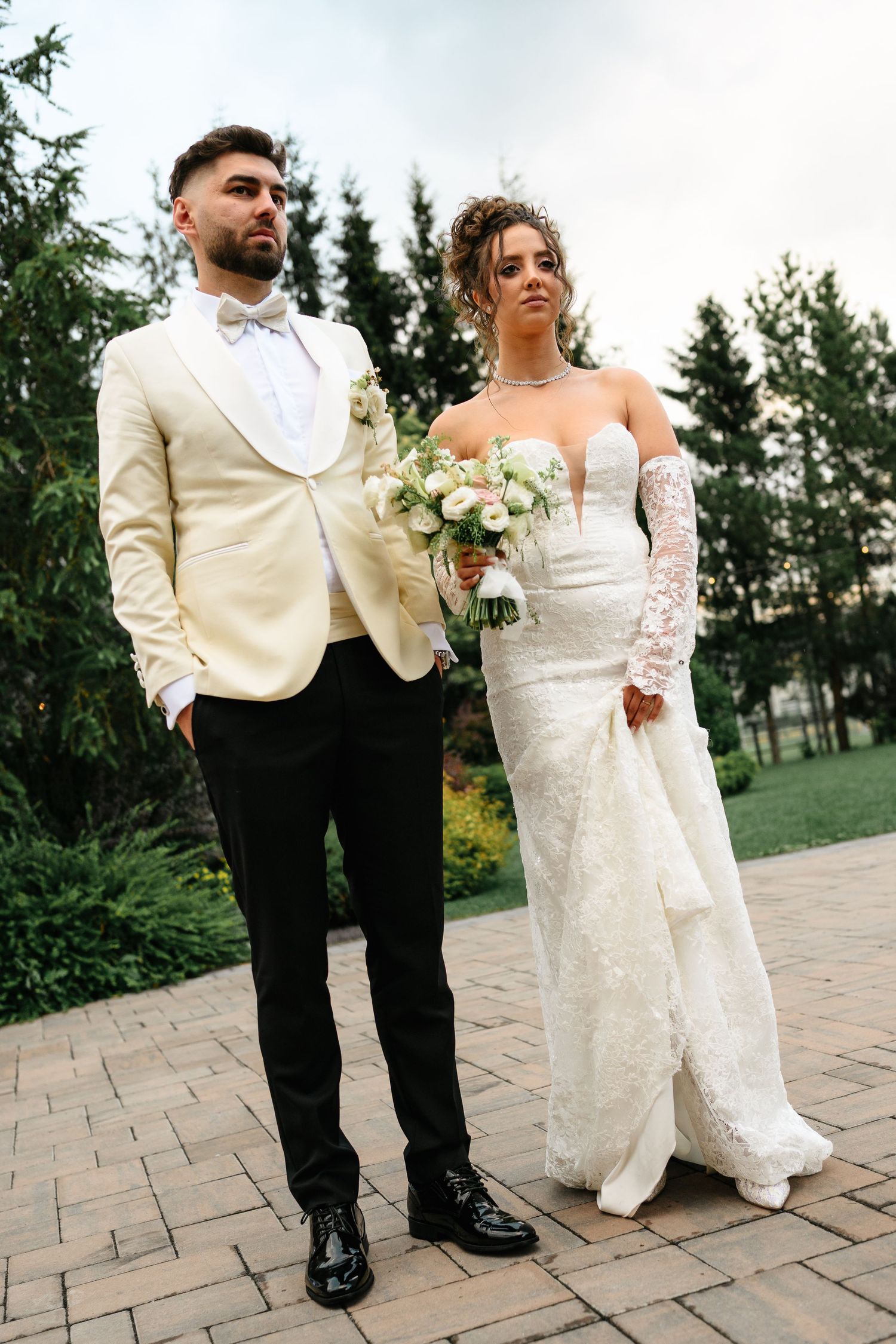 Elegant bride and groom posing outdoors, she in lace gown with bouquet, he in ivory tuxedo jacket with black trousers.
