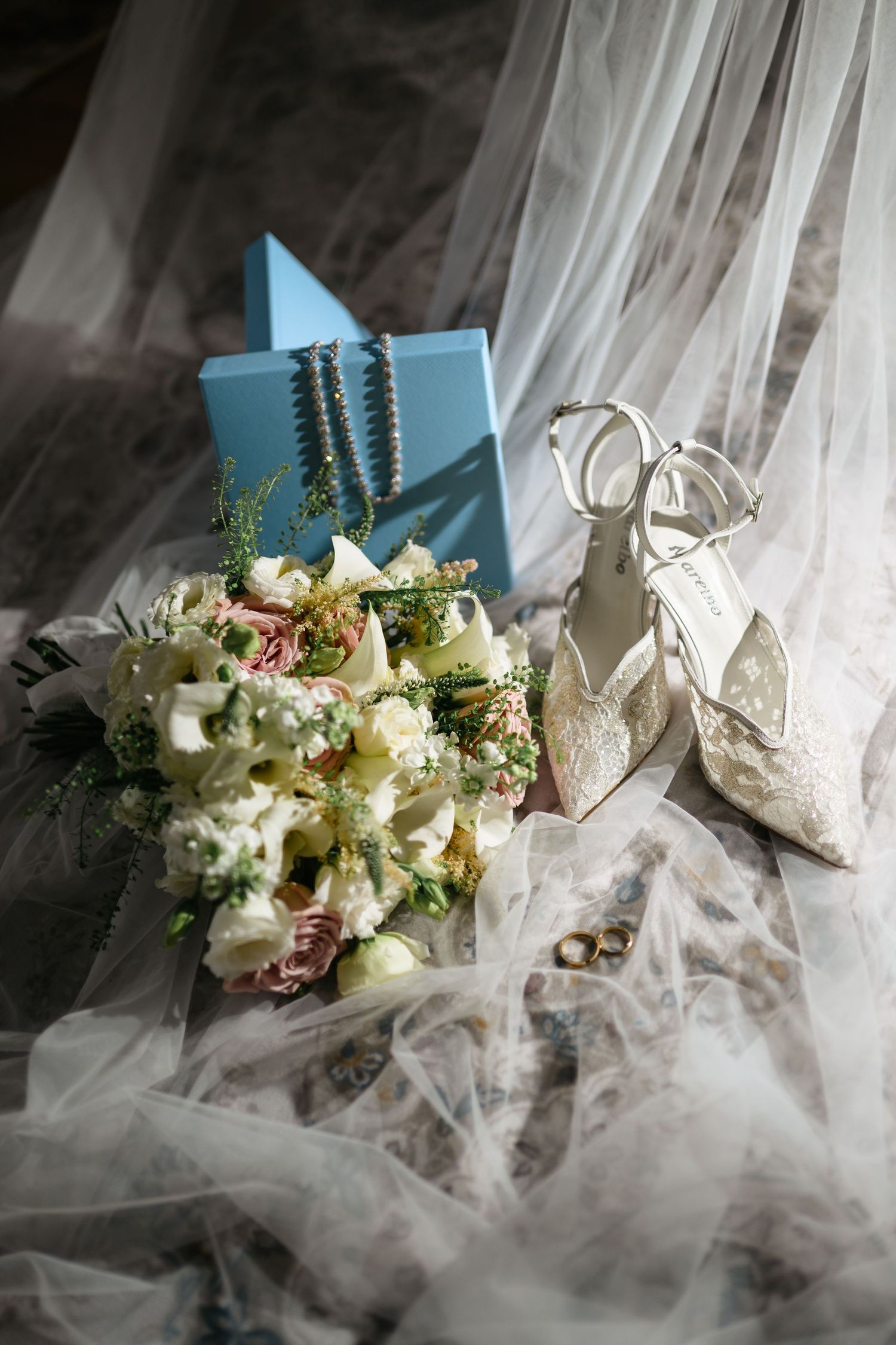 Bridal flat lay with lace heels, floral bouquet, blue gift box, pearl jewelry, and white tulle veil.