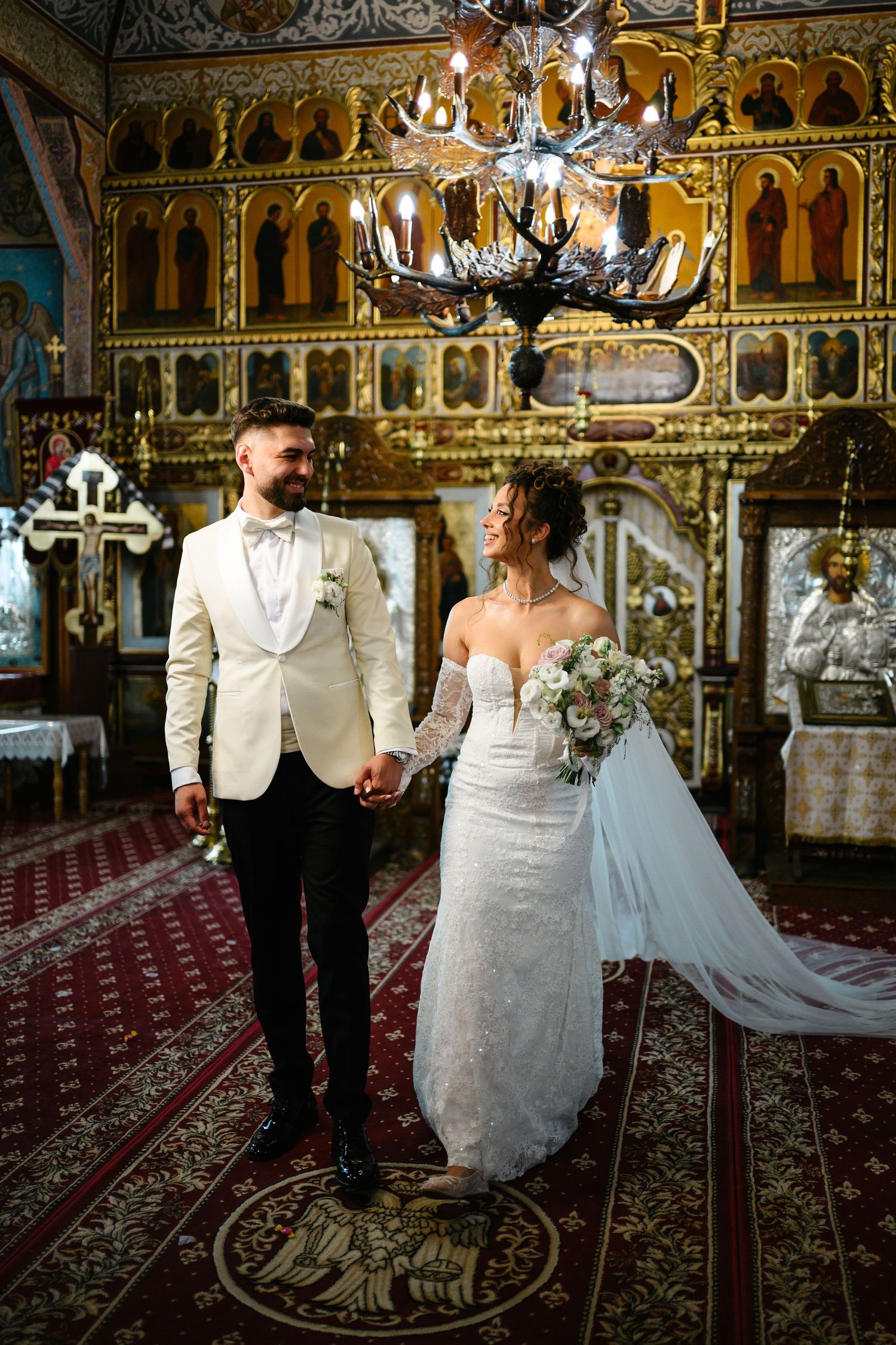 Bride and groom hold hands in ornate Orthodox church with golden iconostasis and chandelier during wedding ceremony.