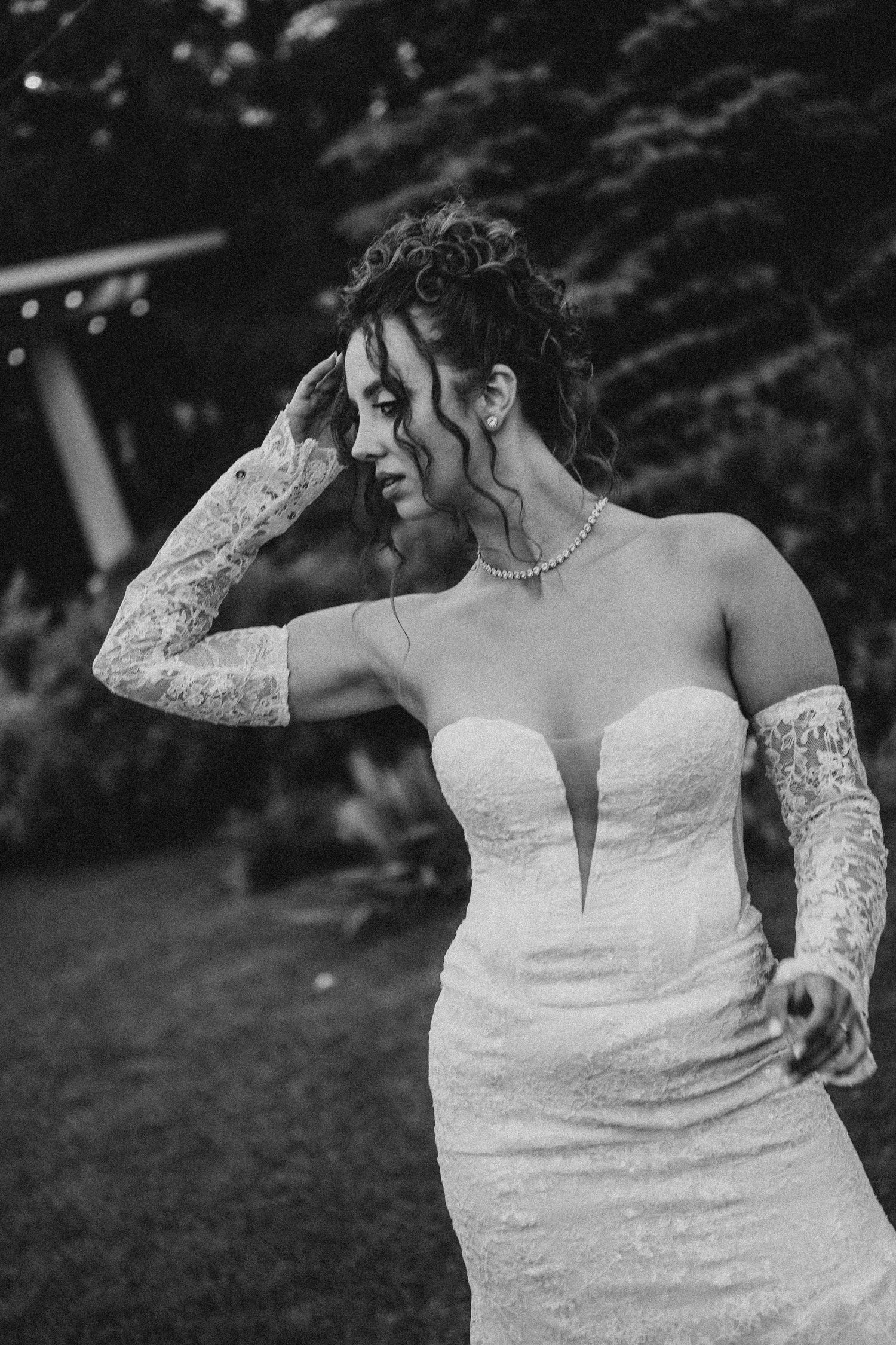Black and white photo of a bride in a strapless lace wedding dress and gloves, posing outdoors at a garden reception.
