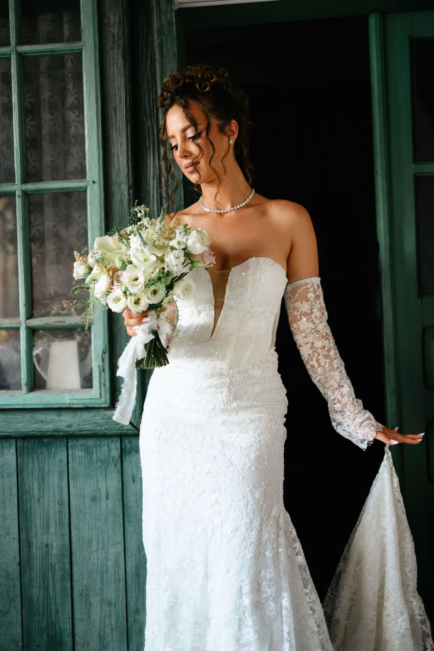 Elegant bride in strapless lace wedding gown with detached sleeves holding white floral bouquet beside green vintage door.
