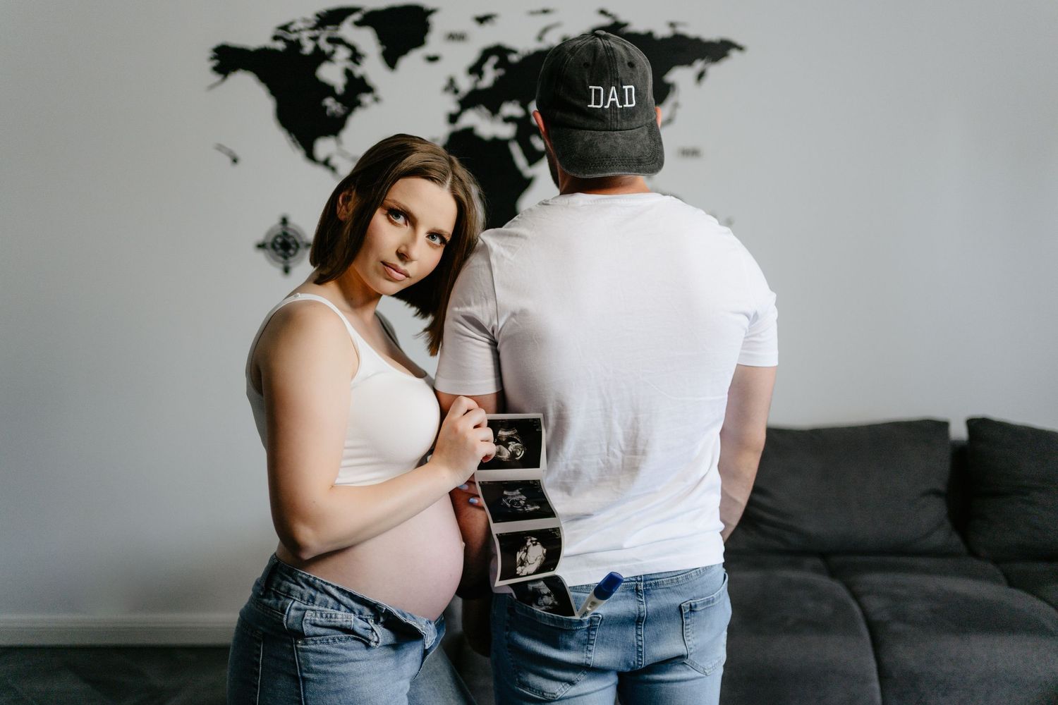 Pregnant woman and man in DAD cap holding ultrasound photos together in a modern living room.