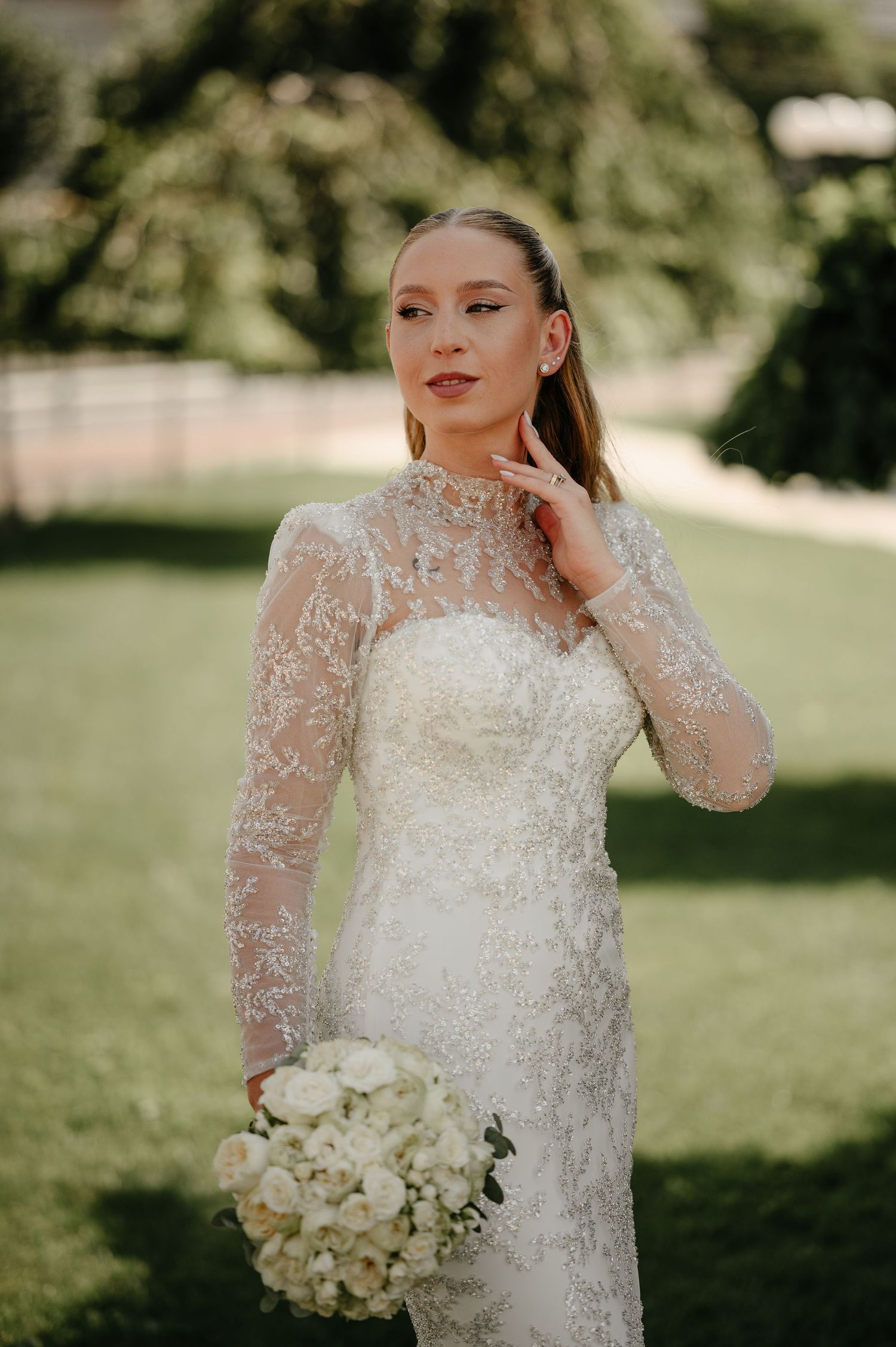 Elegant bride in long-sleeve beaded lace wedding gown holding white roses bouquet in garden.