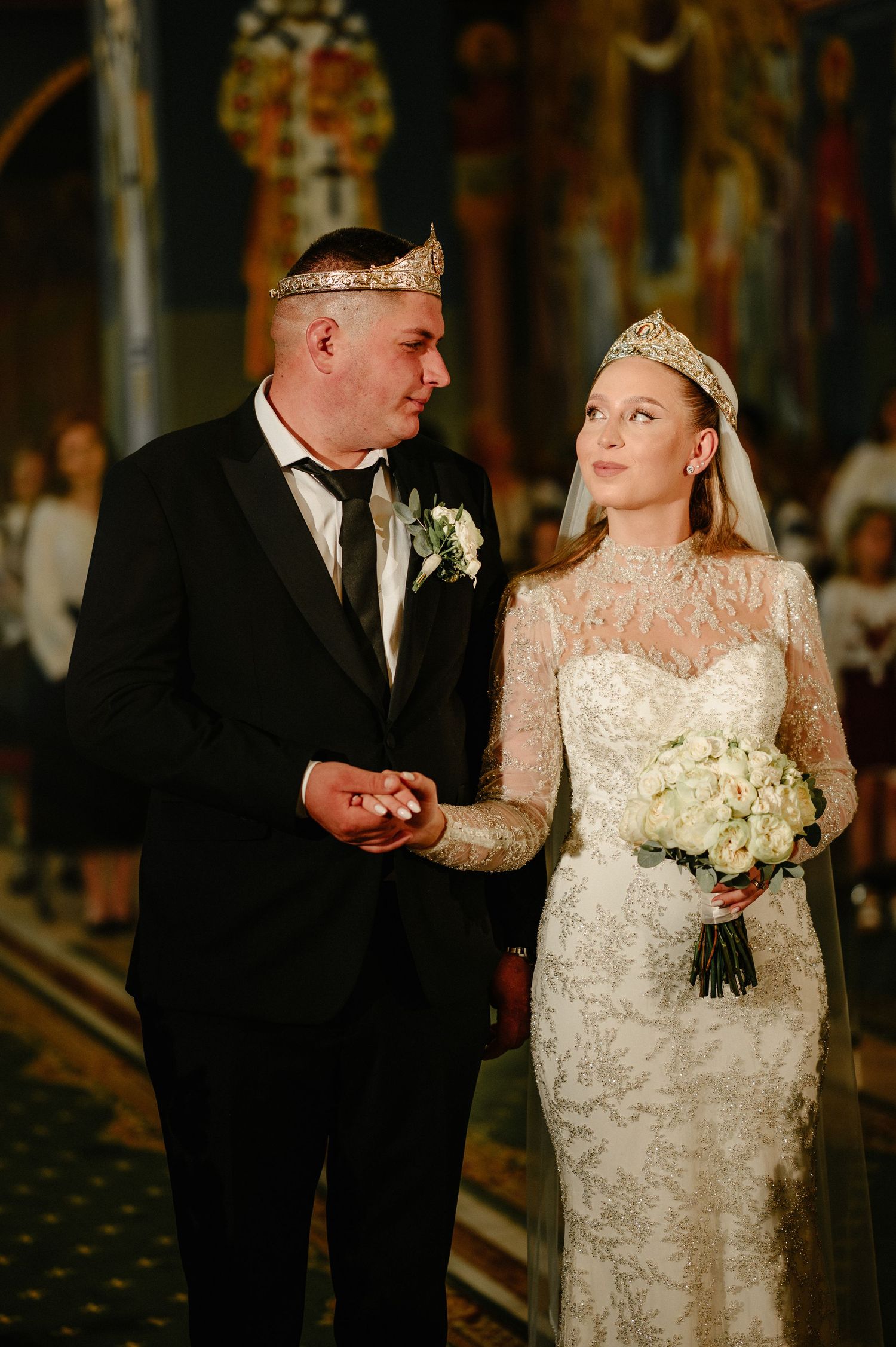 Bride and groom wearing crowns exchange rings during Orthodox wedding ceremony in ornate church.