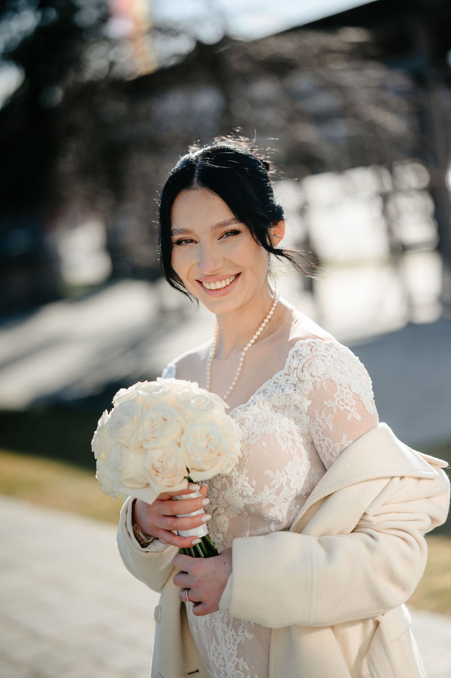 Smiling bride in lace wedding dress, pearl necklace, and beige coat holding white bouquet outdoors.